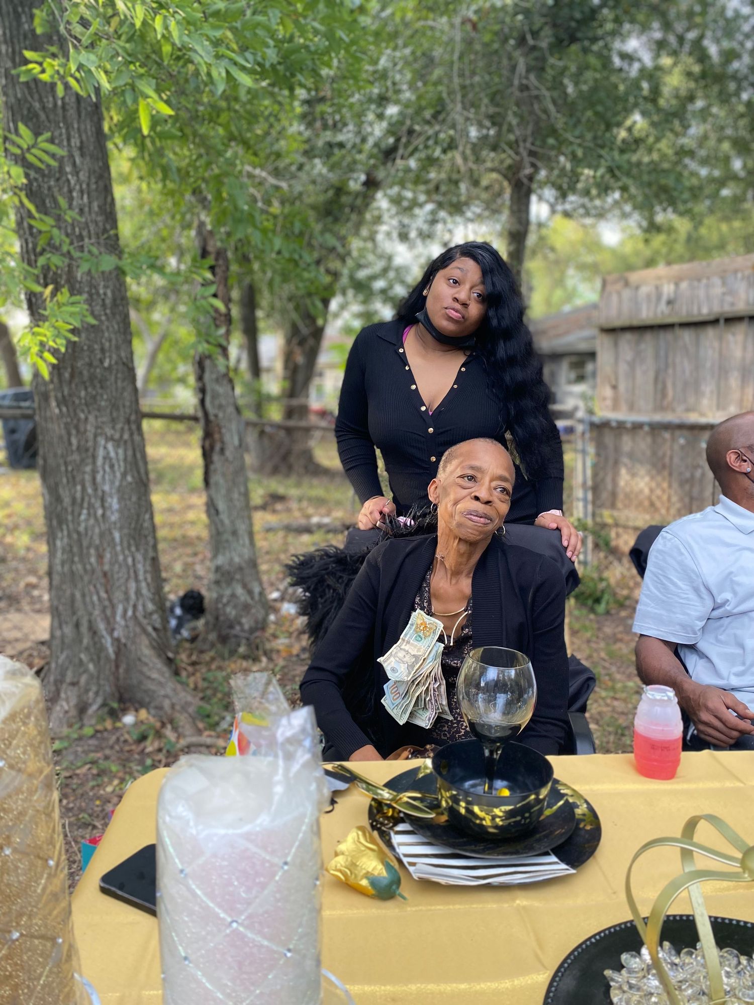 An elderly person seated at a decorated outdoor birthday table with a woman standing behind.