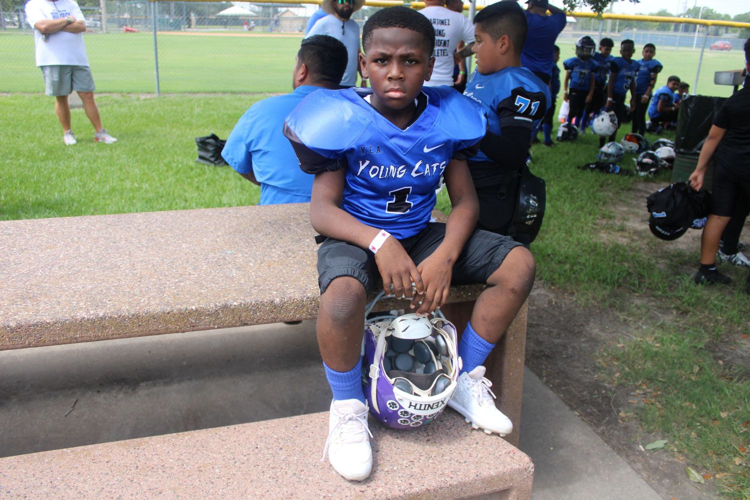 Young boy in blue football uniform sitting on bleachers holding helmet during youth football practice.