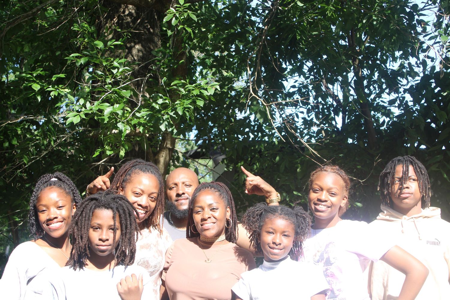 A group of children and an adult posing together outdoors under a large green tree.