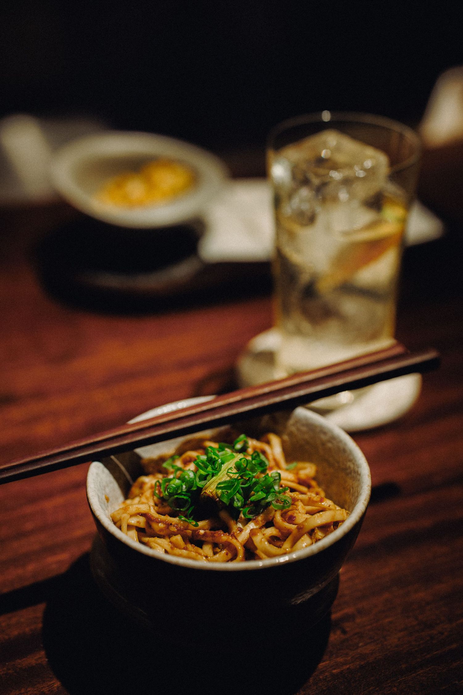 Bowl of Asian noodles topped with herbs and chopsticks resting on rim beside an iced drink.
