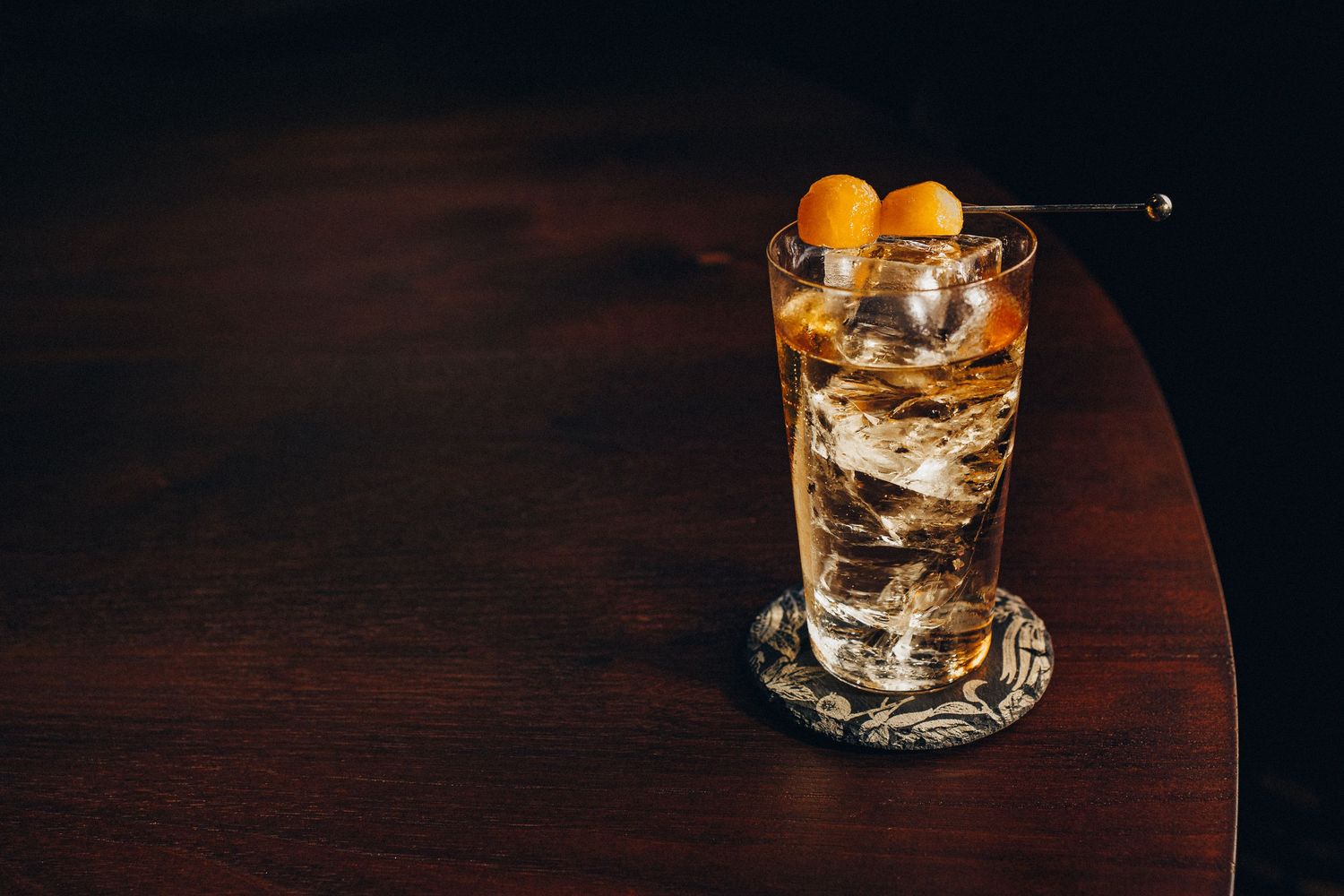 Classic whisky highball cocktail with ice and orange cherry garnish on dark wooden table.