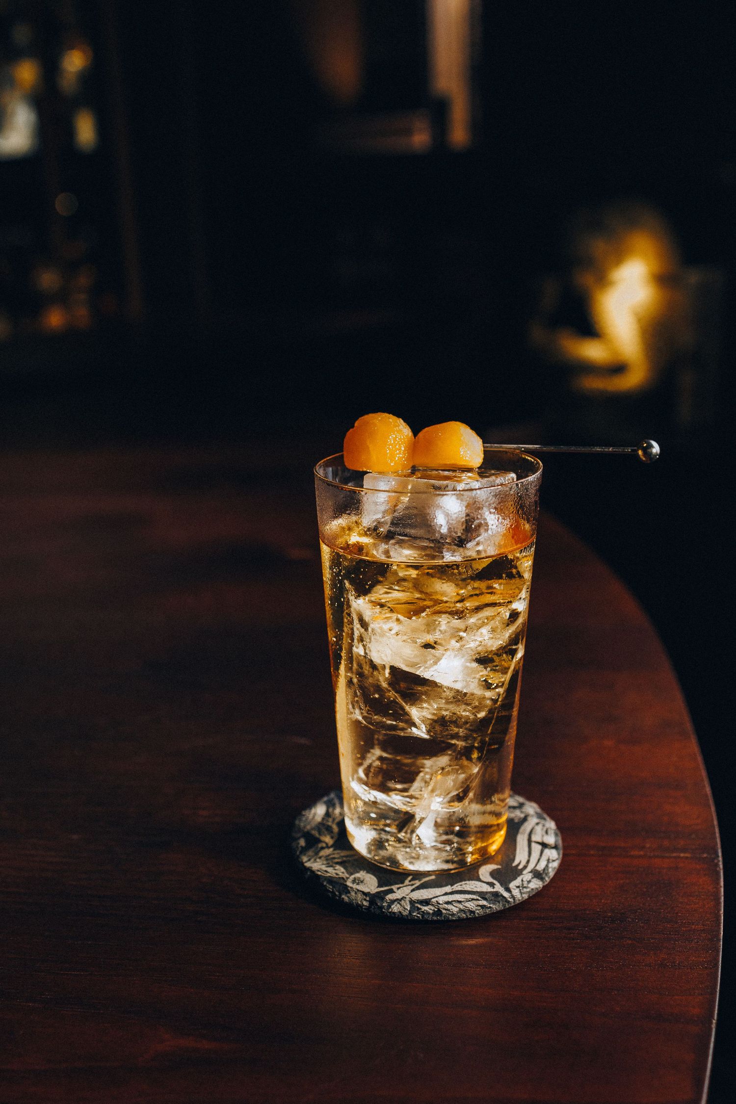 Whiskey highball cocktail with ice and melon garnish on a decorative coaster at a bar.