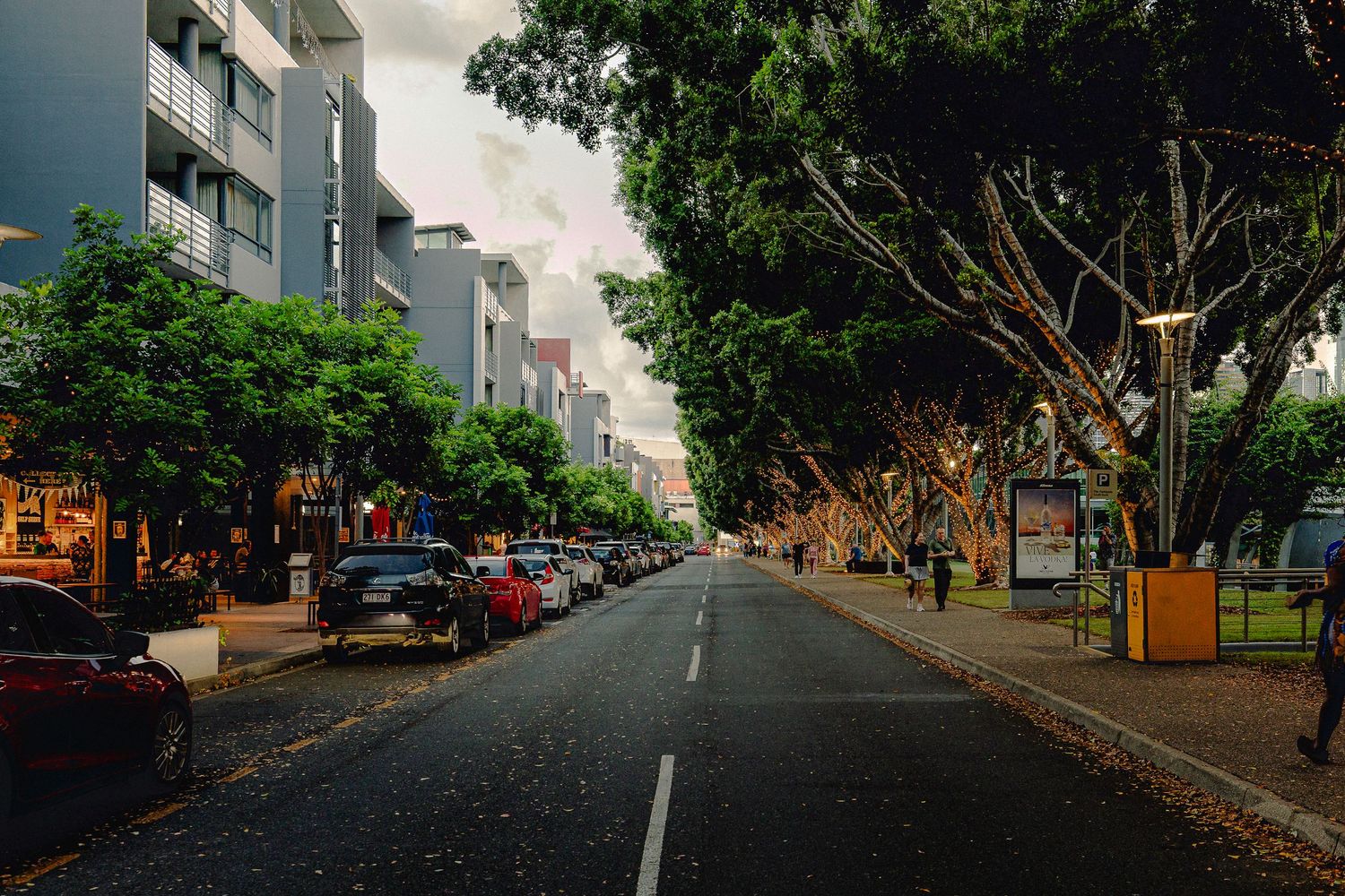 A tree-lined urban street with parked cars and modern buildings under a cloudy sky.