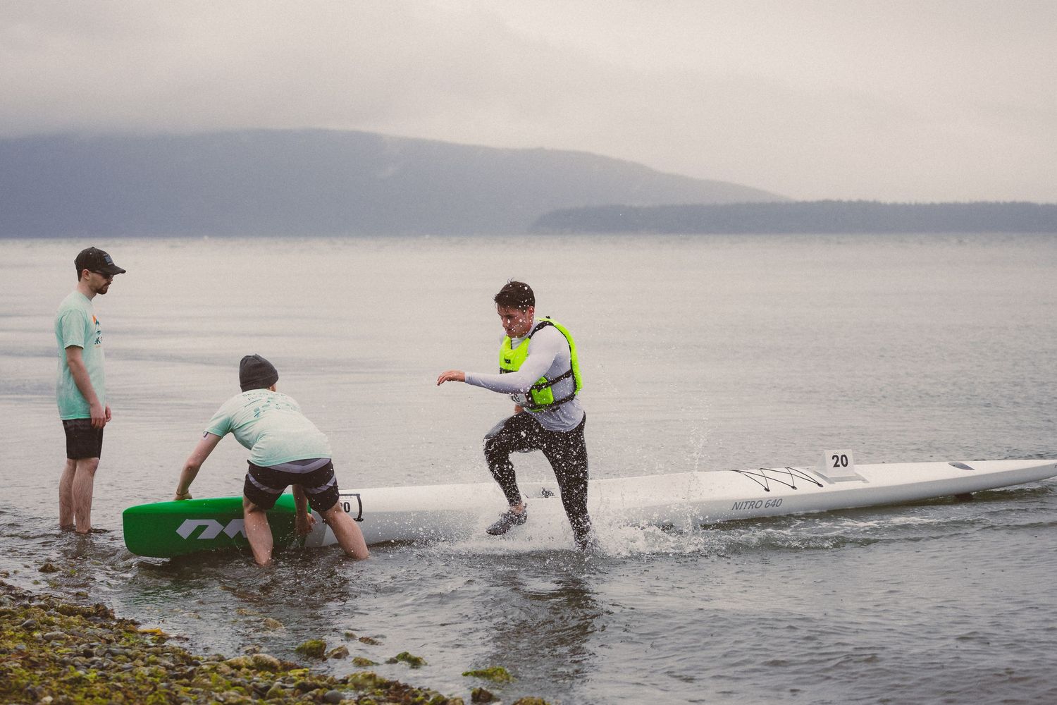 Athletes help a kayaker exit a white kayak onto a rocky shoreline during a triathlon transition.