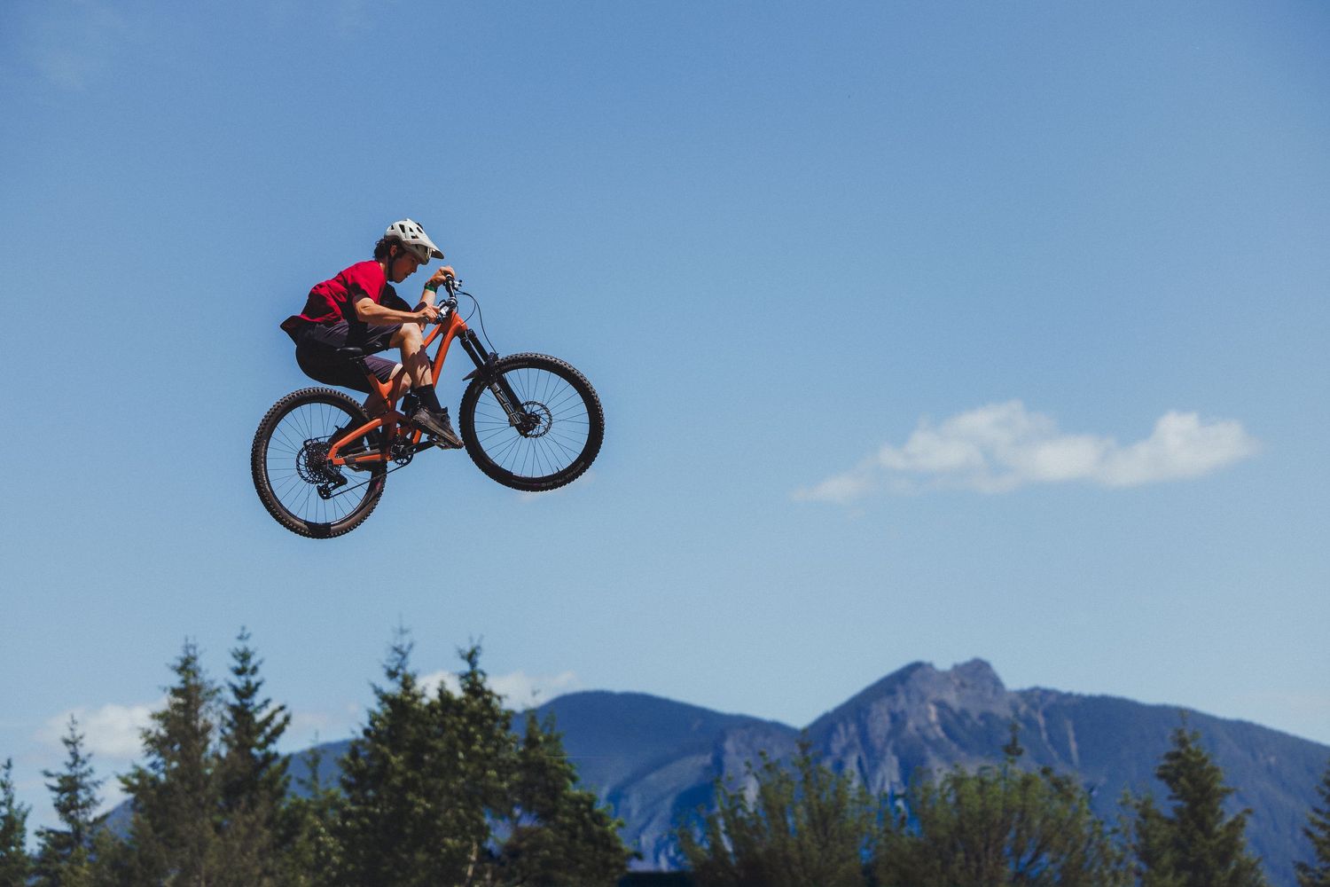 Mountain biker catches big air on a jump with pine trees and a scenic mountain peak in the background.