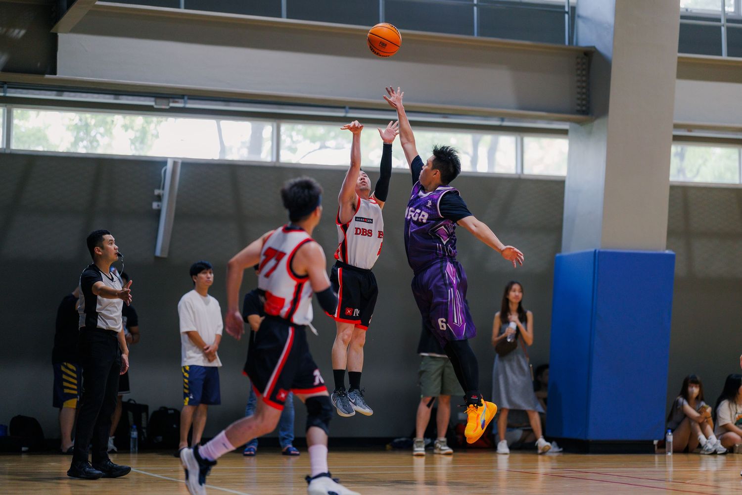 Basketball players competing for a jump ball during an indoor game with spectators watching from the sideline.