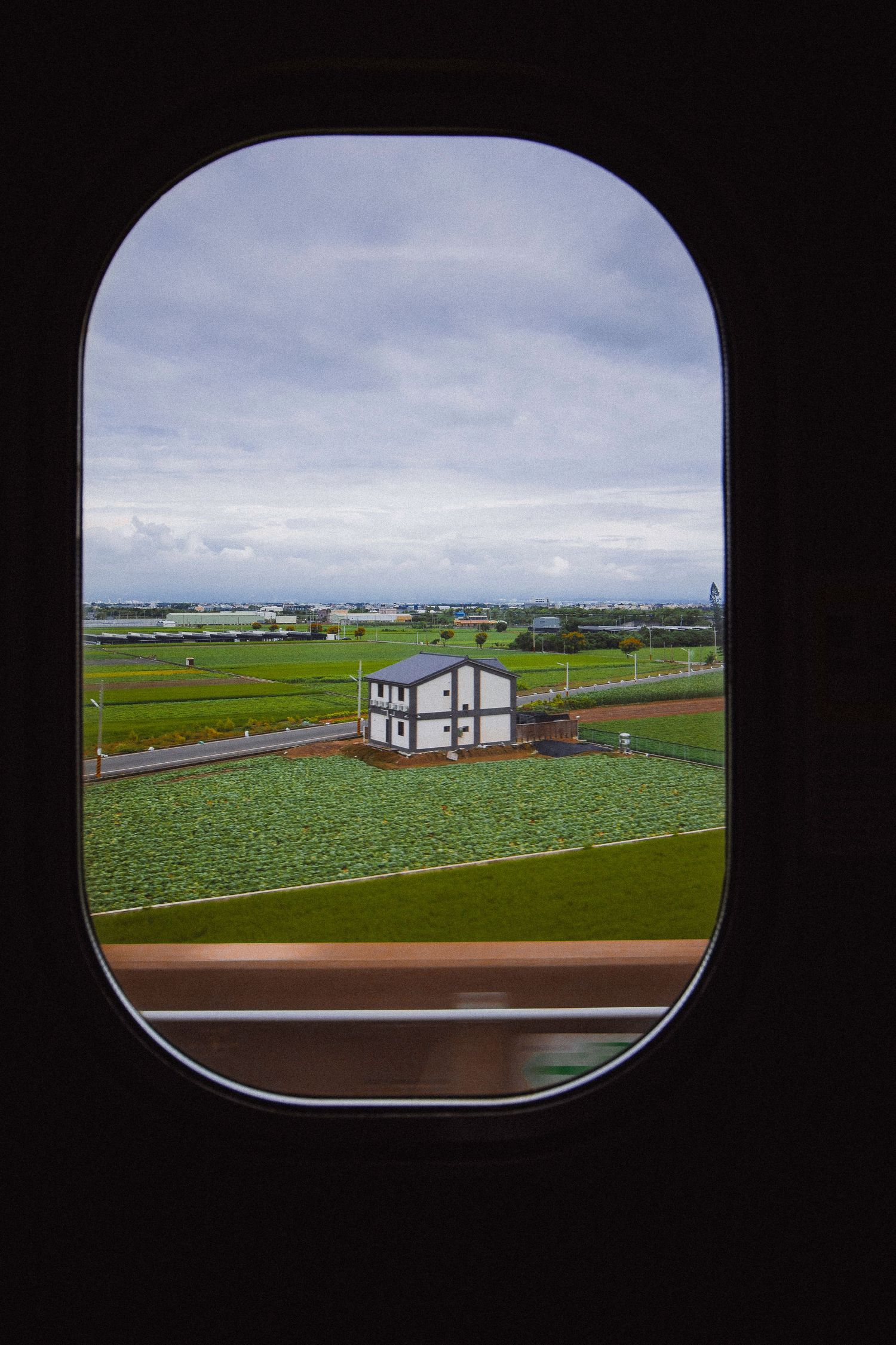 View through airplane oval window of a small white farmhouse surrounded by green fields under cloudy sky.