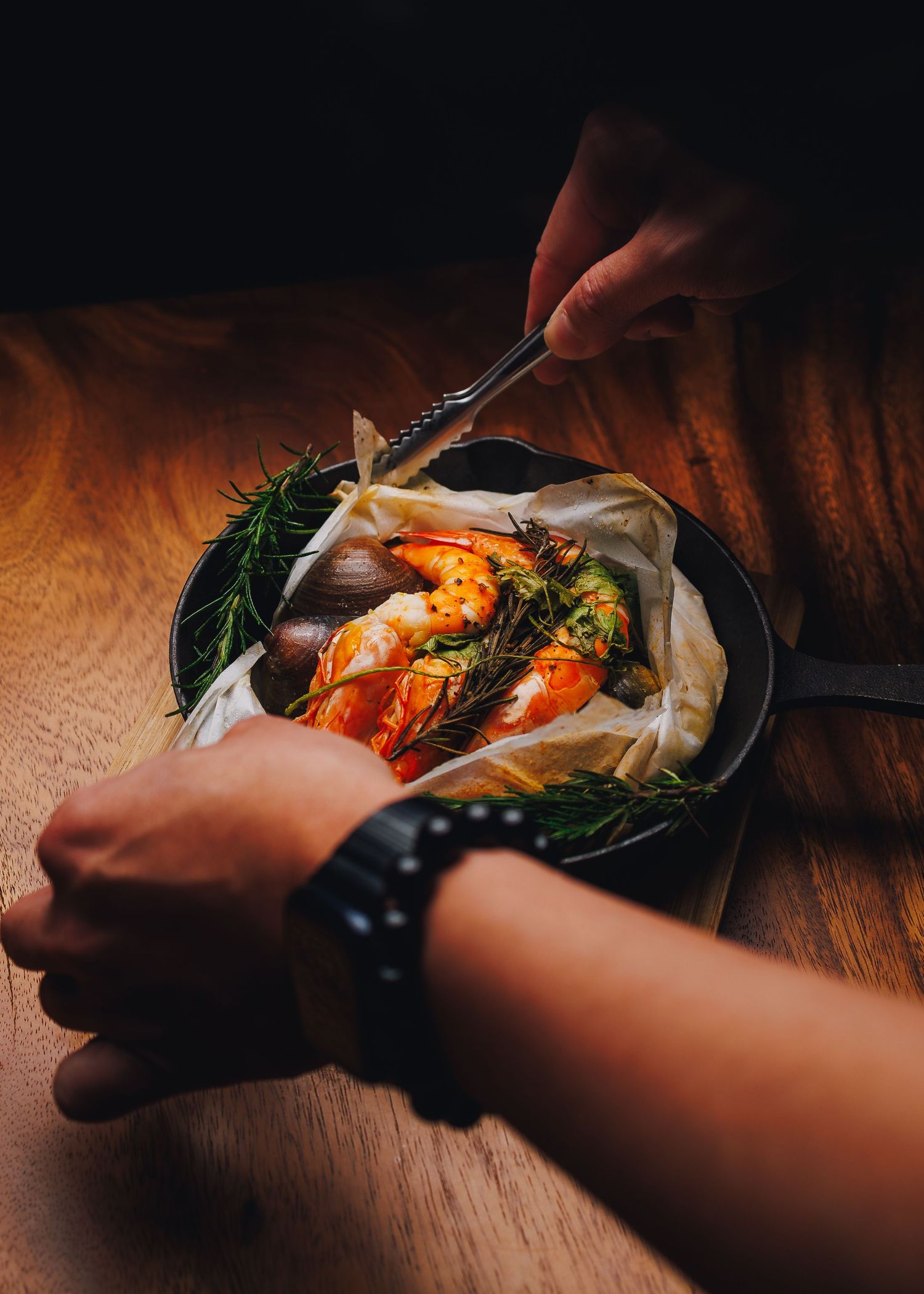 Hands using tongs to serve shrimp and vegetables in a rustic cast iron skillet.