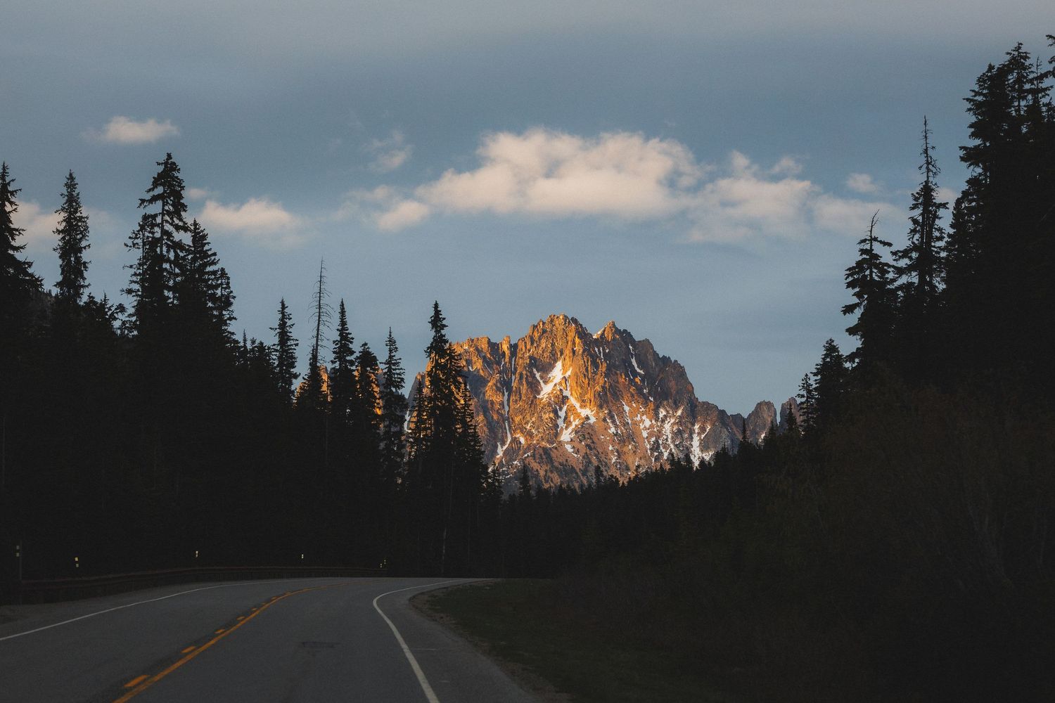 Sunlit rocky mountain peak at dusk framed by tall pine trees along empty highway.