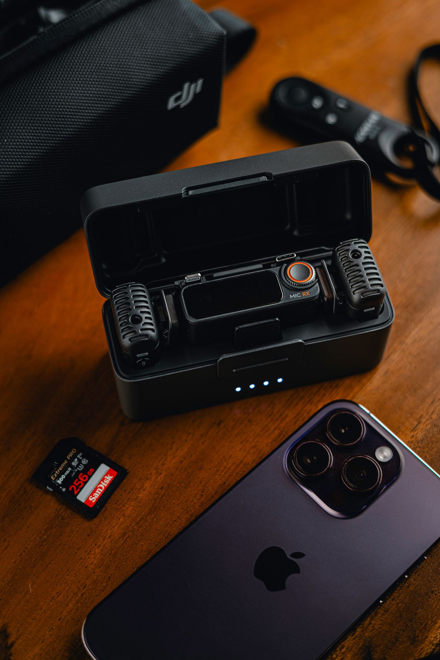 DJI wireless microphone system in charging case next to iPhone 14 Pro and SanDisk SD card on wooden desk.