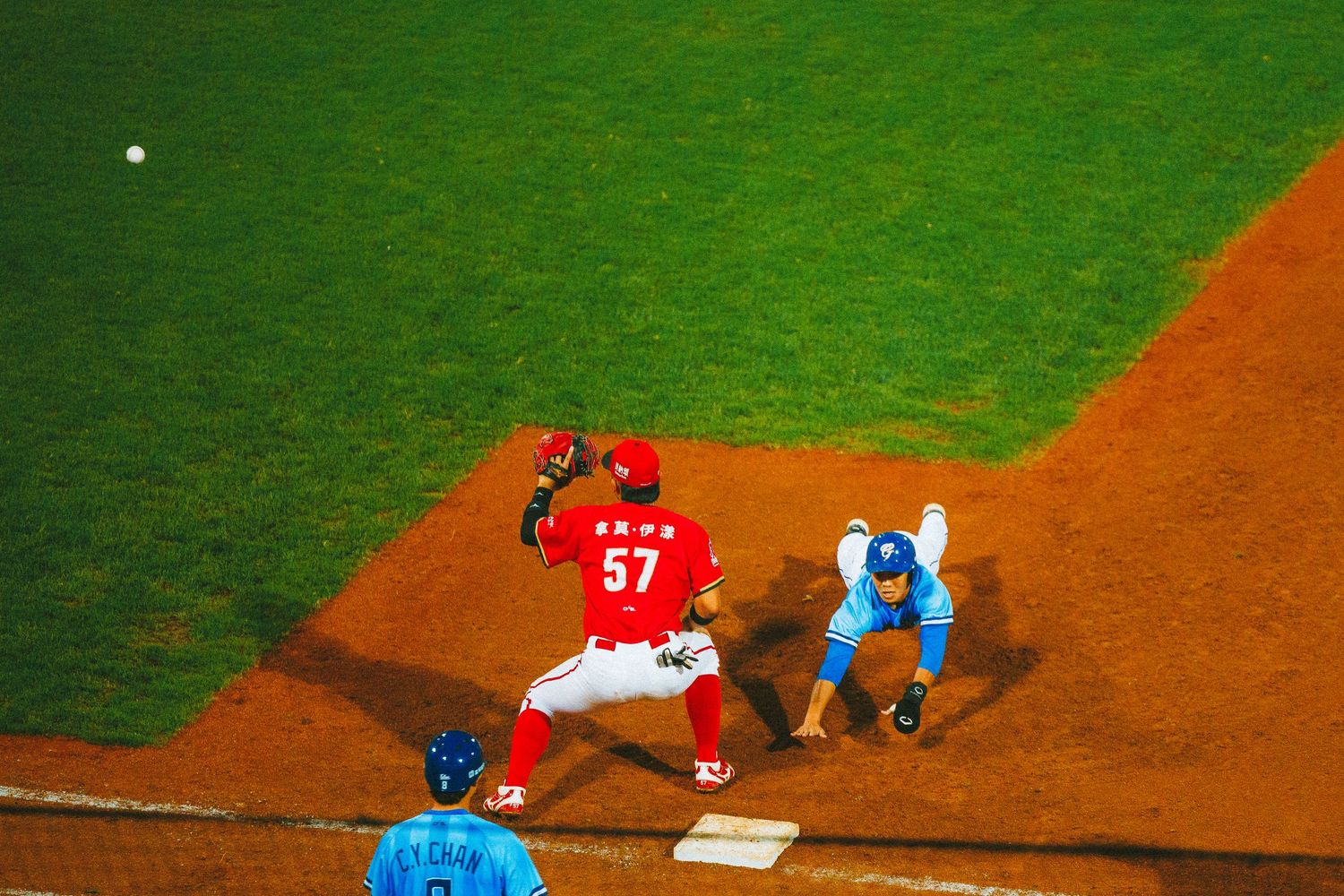 Baseball first baseman in red uniform #57 catching a throw as a runner slides into first base during a game.
