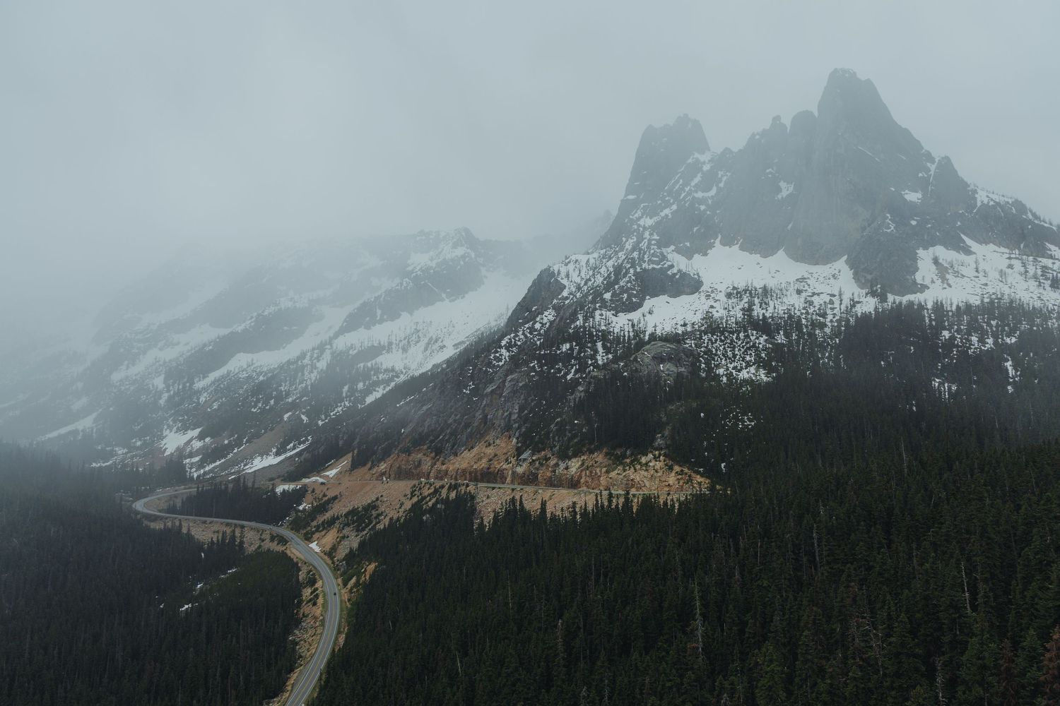Snow-capped mountain peaks shrouded in fog above dense pine forest and winding road.