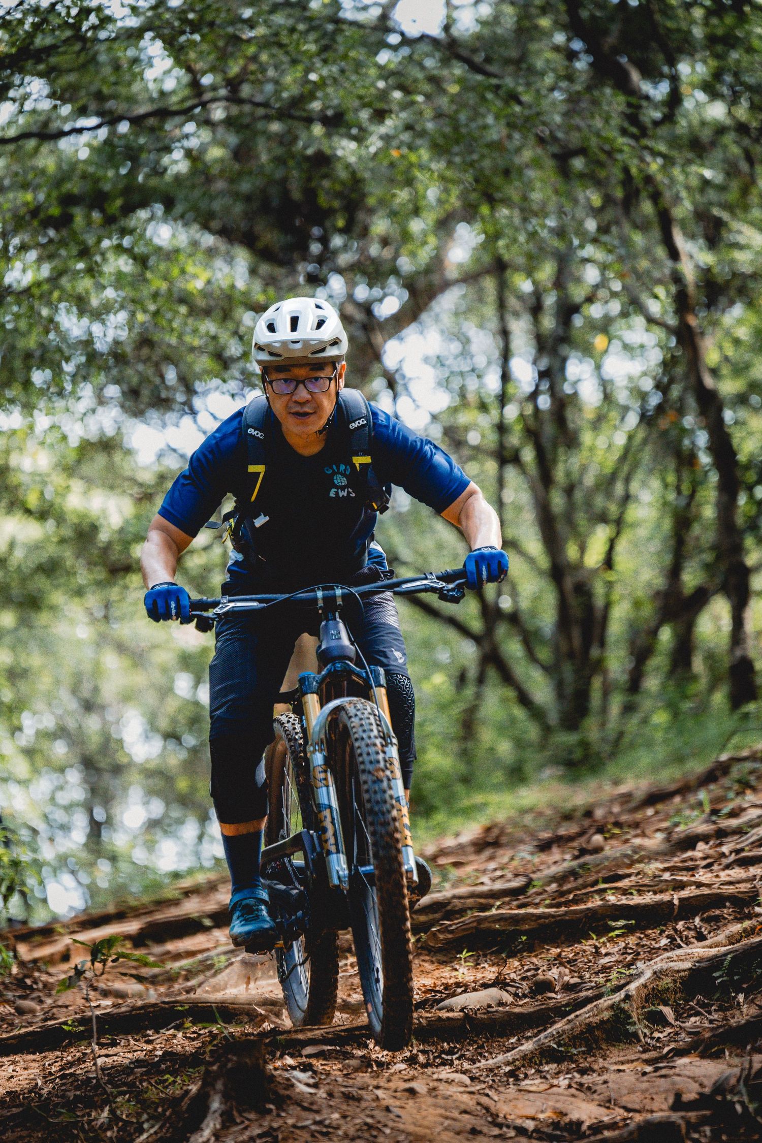 Mountain biker wearing white helmet and blue jersey number 7 riding downhill on a forest trail.