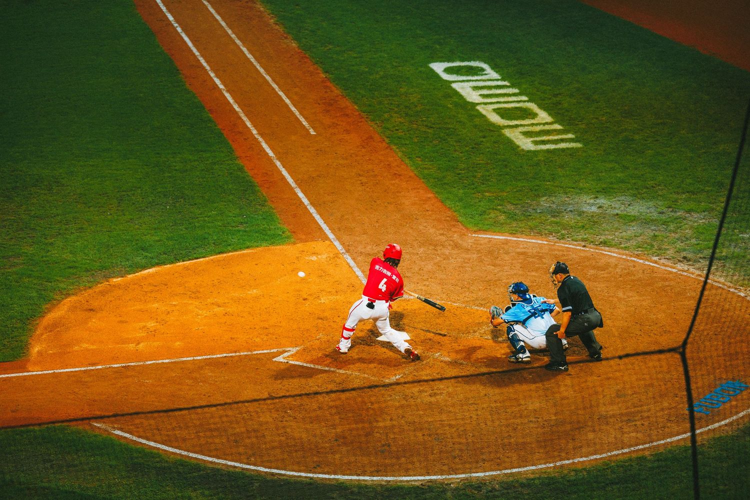 A baseball batter in red swings at a pitch during a professional stadium game with umpire present.