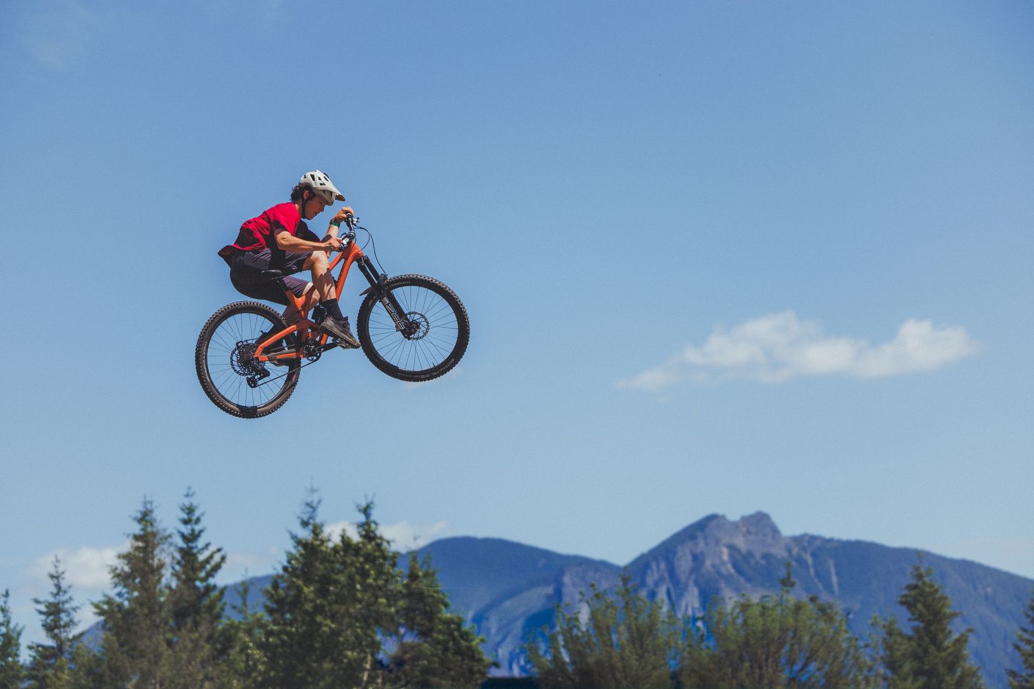Mountain biker performing high aerial jump trick against clear blue sky with mountain backdrop.