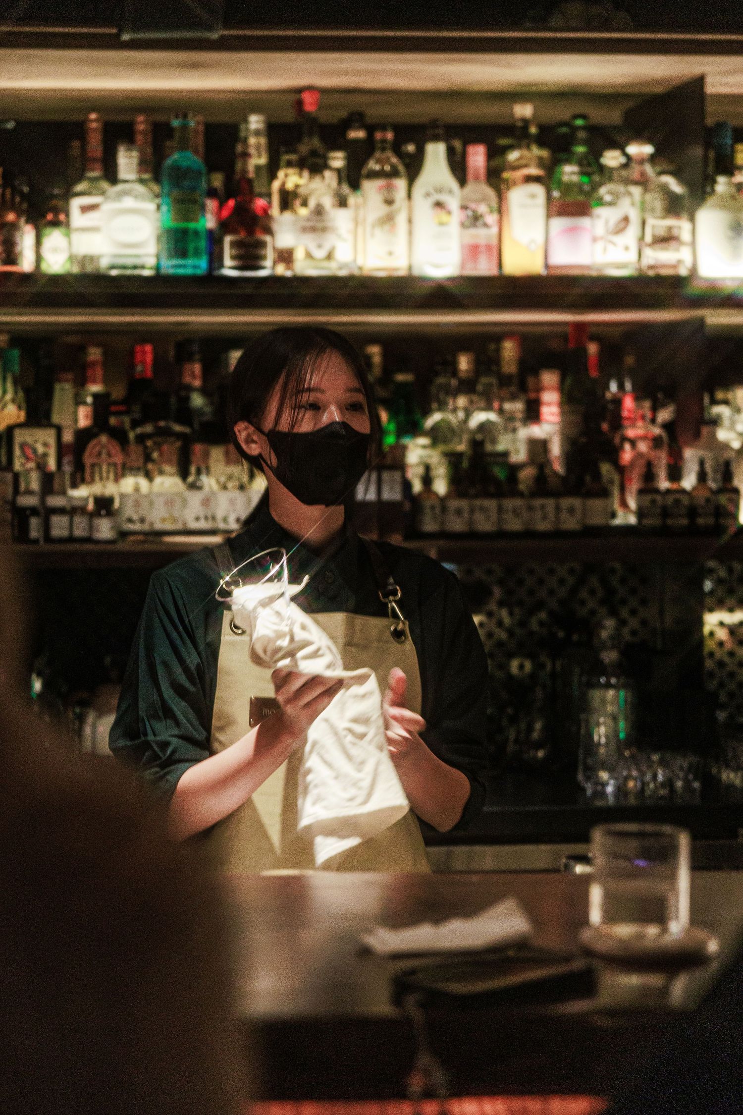 Female bartender in black mask and apron working behind a well-stocked cocktail bar counter.