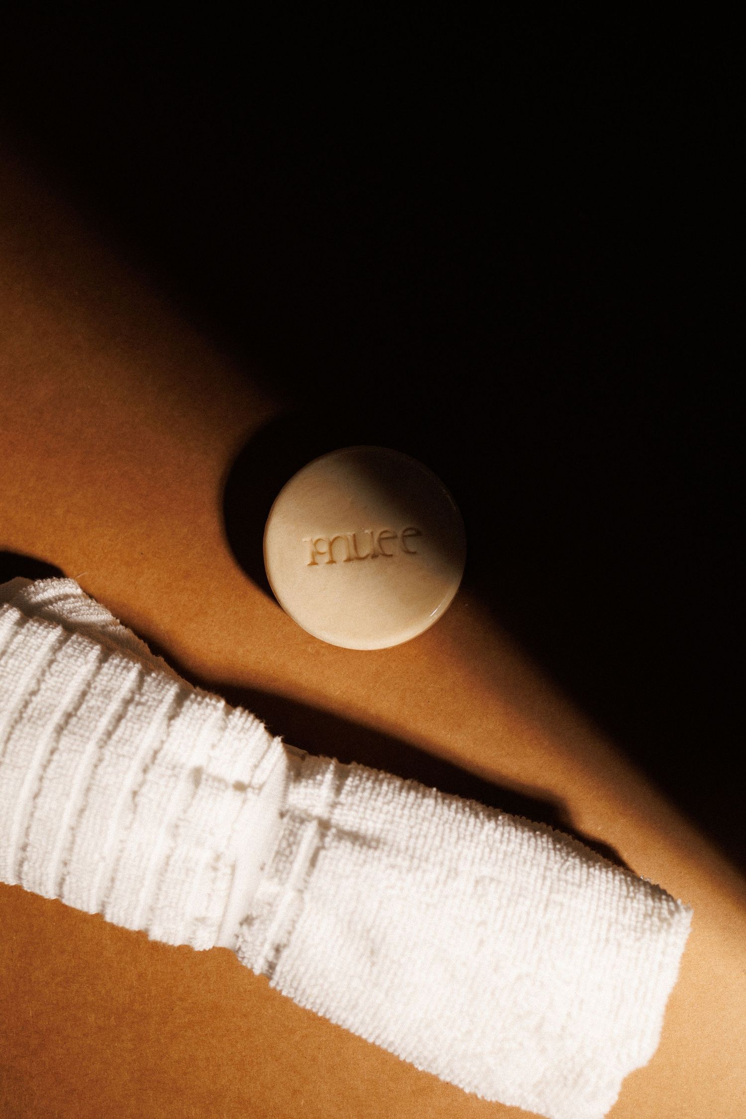 Muee branded round soap bar resting on a white washcloth in dramatic shadow lighting.