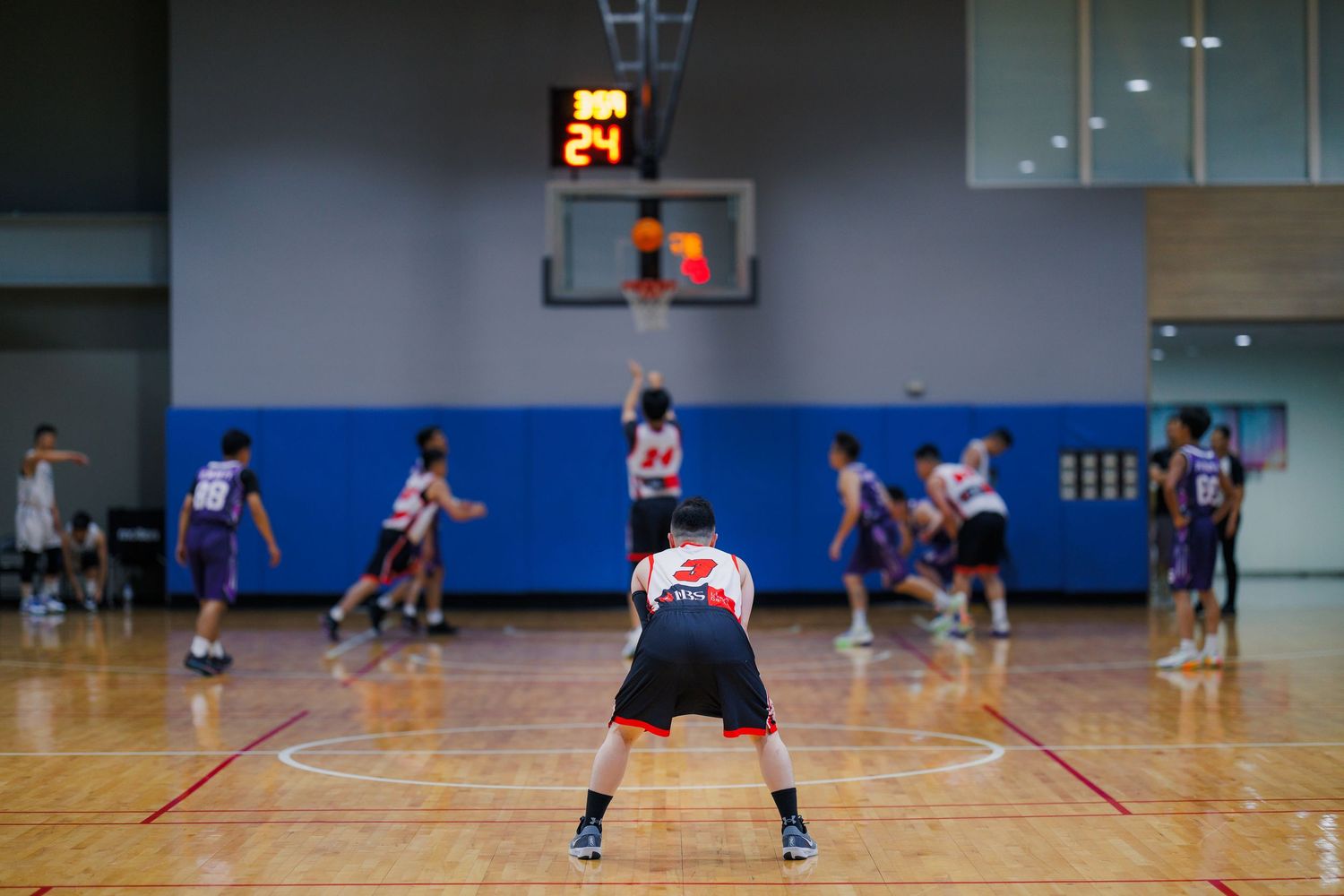 Basketball player crouching in defensive stance on indoor court during competitive game with shot clock visible.