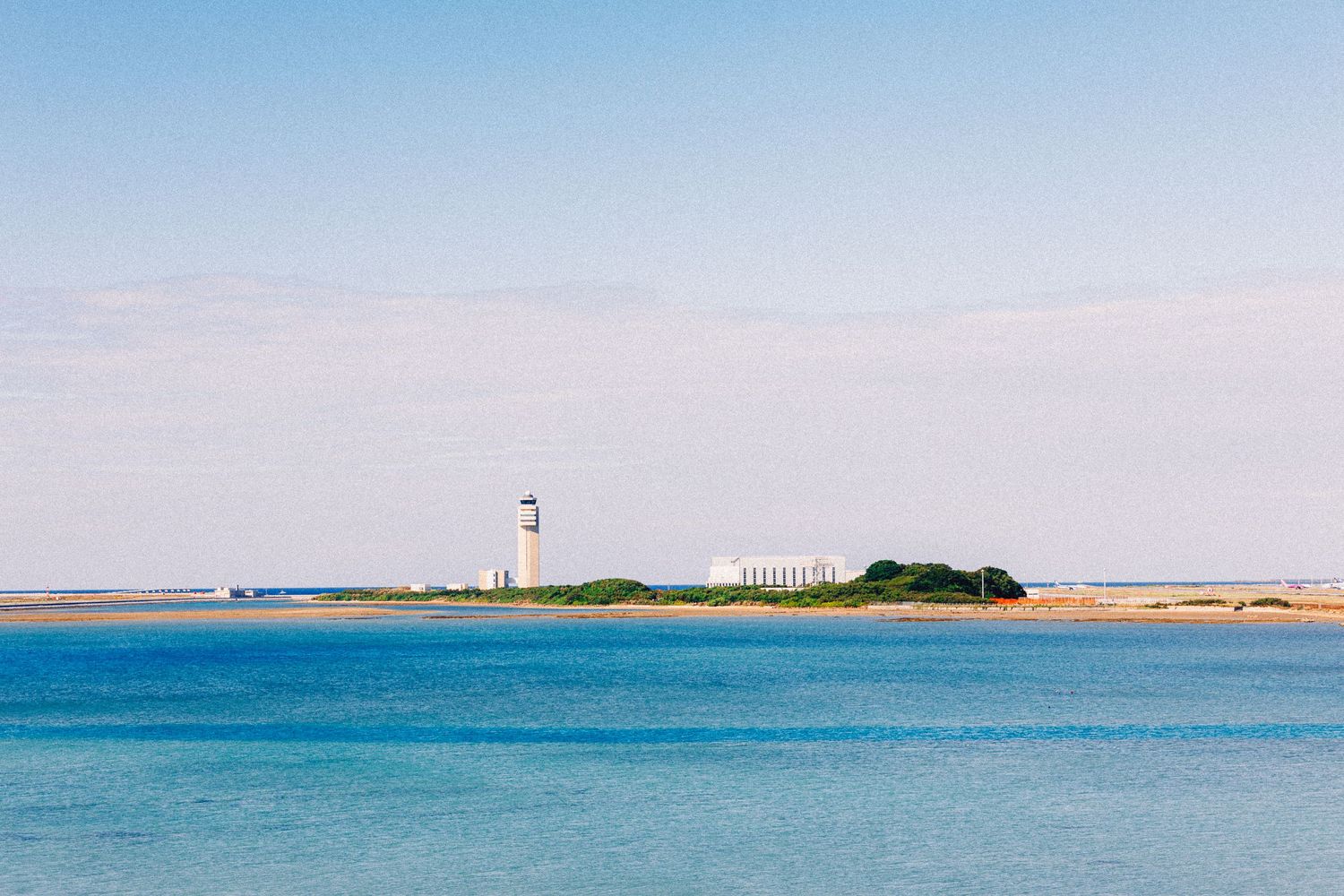 A lighthouse stands on a sandy island surrounded by calm turquoise waters under a clear blue sky.