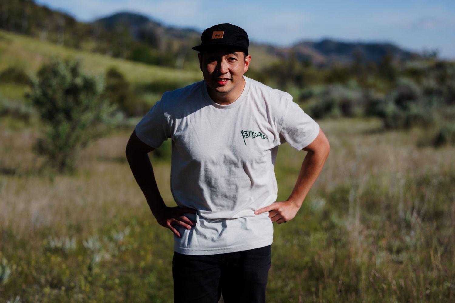 A man in a white t-shirt and black cap stands with hands on hips in an open grassy field.
