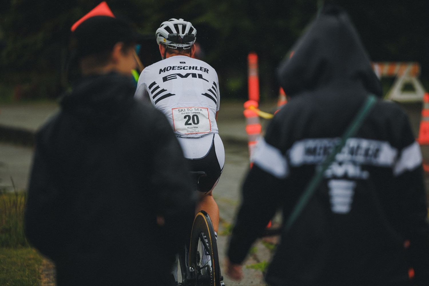 Cyclist wearing number 20 in Evil Bikes kit rides away from support crew in dark hoodies at a race.