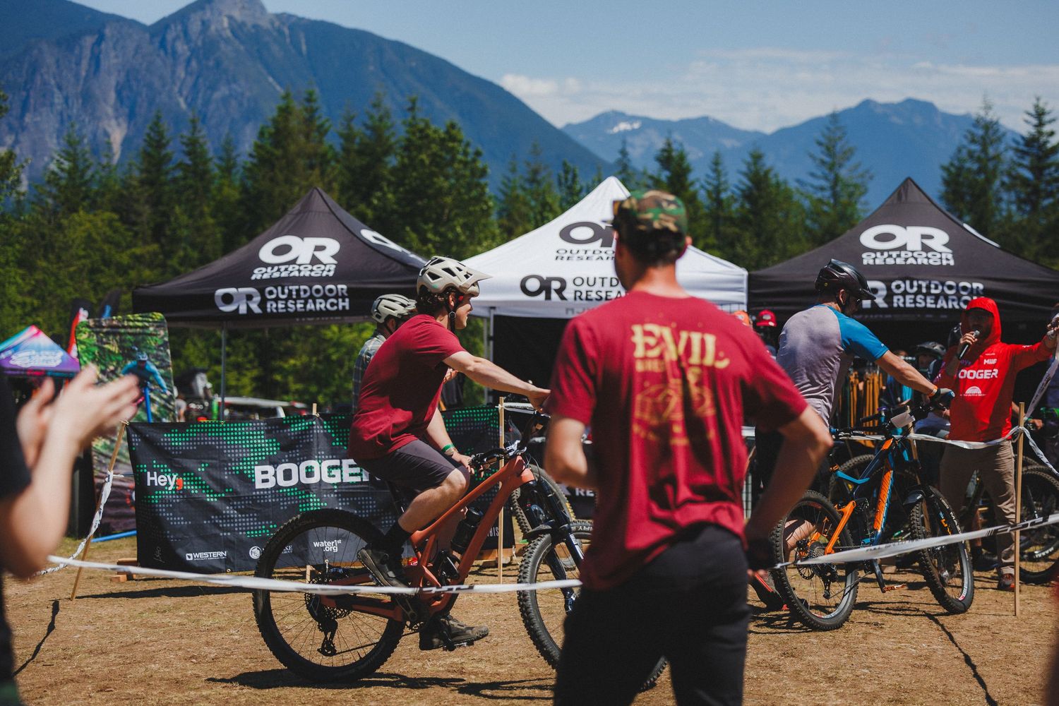 Mountain bikers gather at an Evil Bikes outdoor event with Outdoor Research sponsor tents and mountain views.