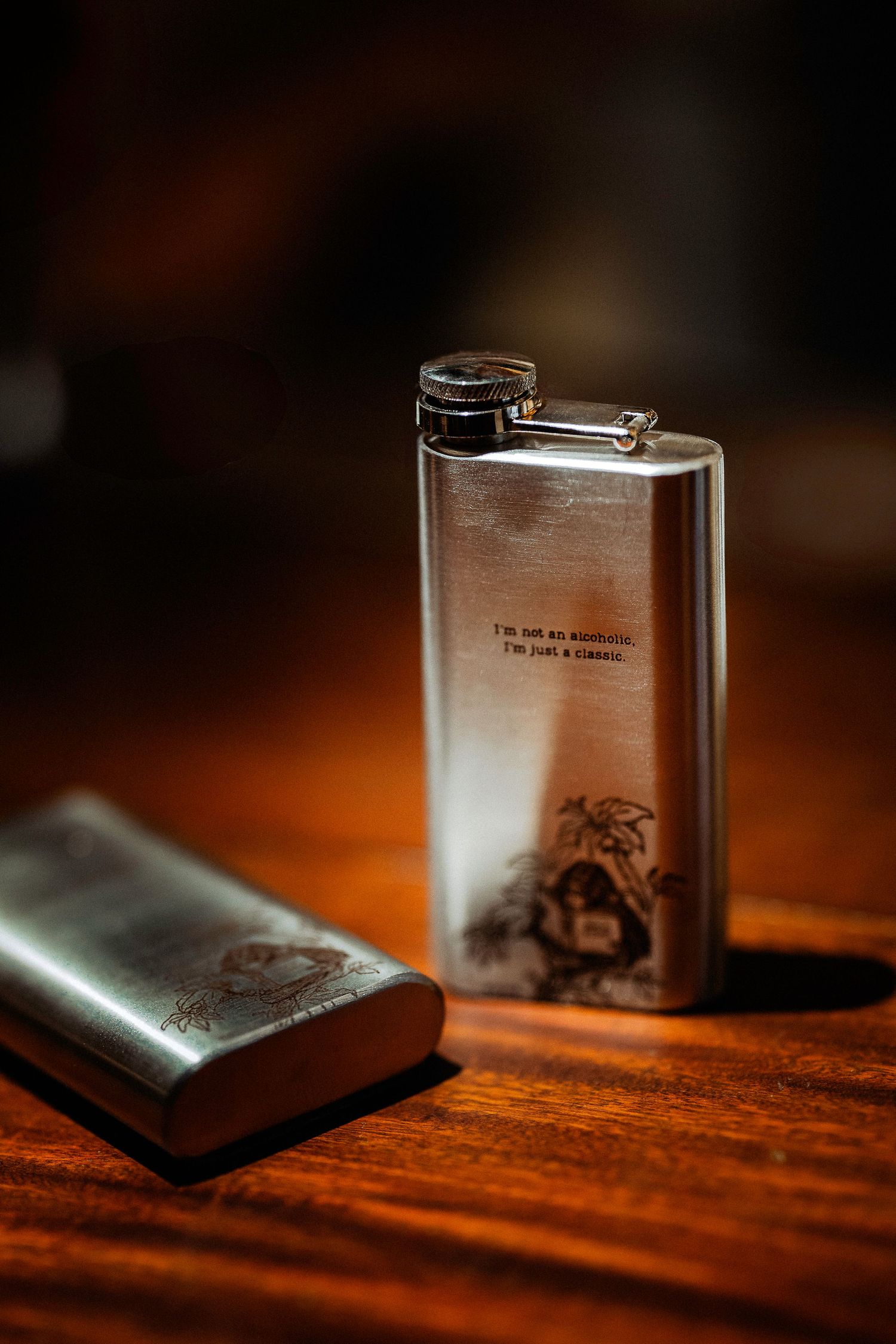 Engraved stainless steel hip flask with text reading I'm not an alcoholic, I'm just a classic on wooden table.