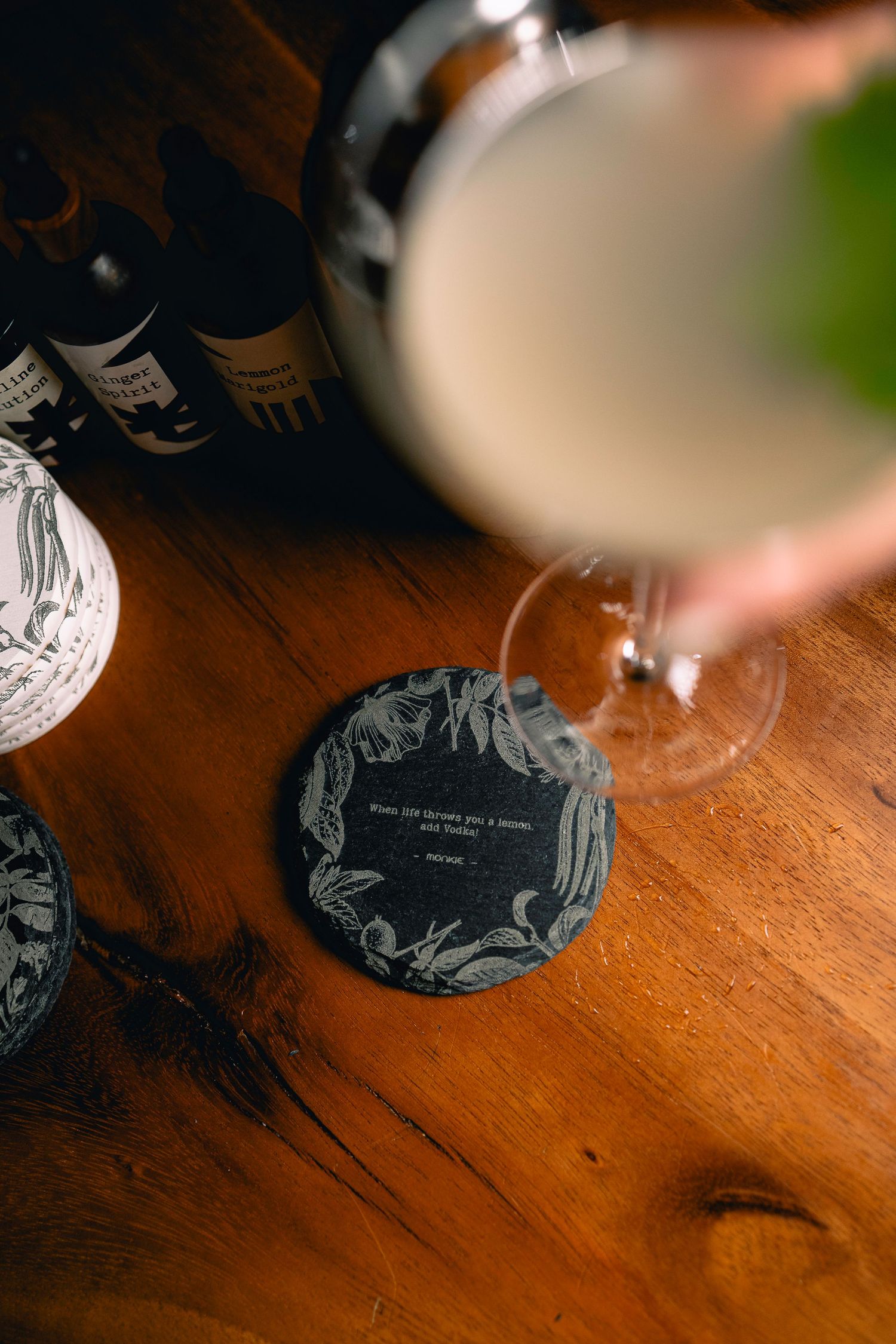 Dark slate coaster with floral engraving beside a wine glass and bottles on wood table.