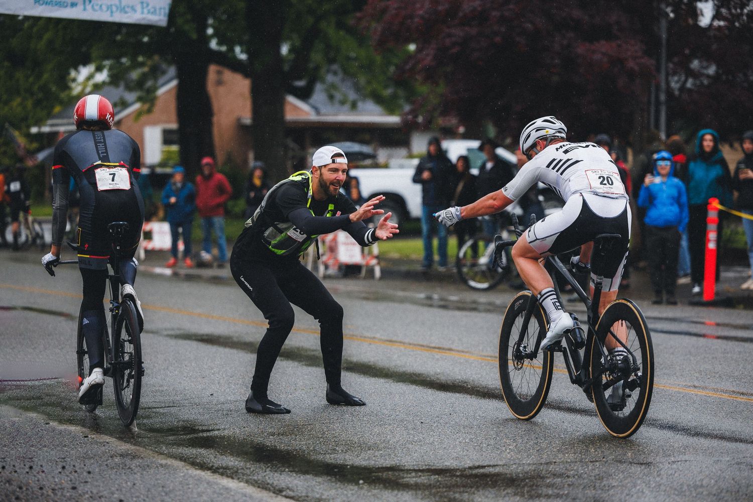 Cyclists compete in a rainy outdoor road race while a coach gestures instructions from the sideline.