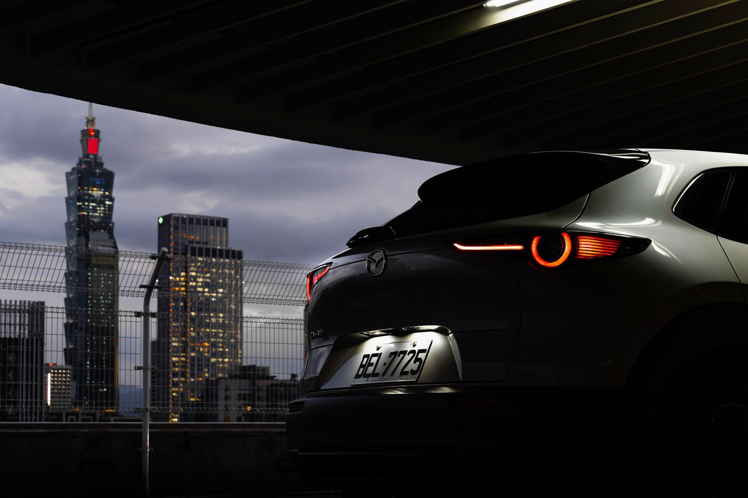 Mazda hatchback rear view with glowing taillights against Taipei 101 skyline at dusk.