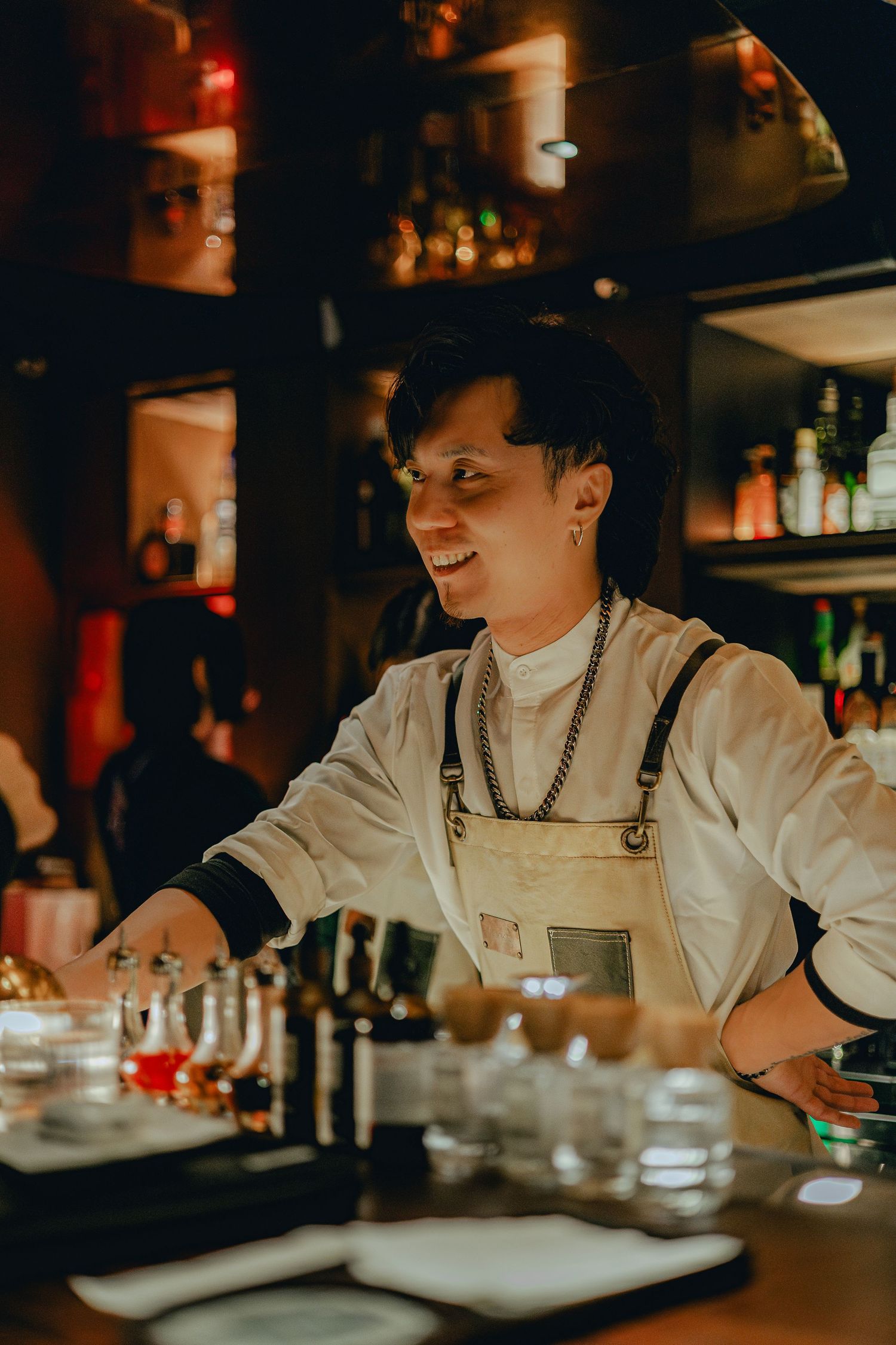 Smiling Asian bartender in white apron arranging glasses on illuminated bar counter at night.