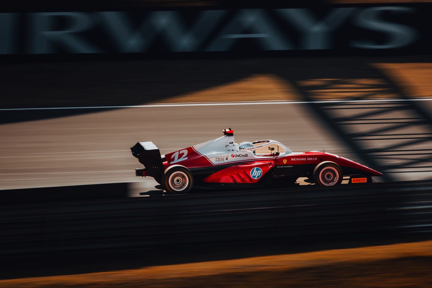 Red Formula 3 car number 22 racing at speed on track in golden sunlight.
