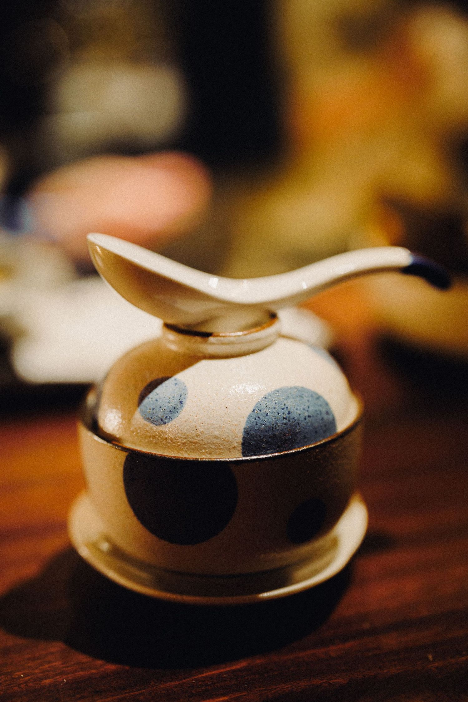 Ceramic polka dot bowl with lid and decorative spoon balanced on top on a wooden table.