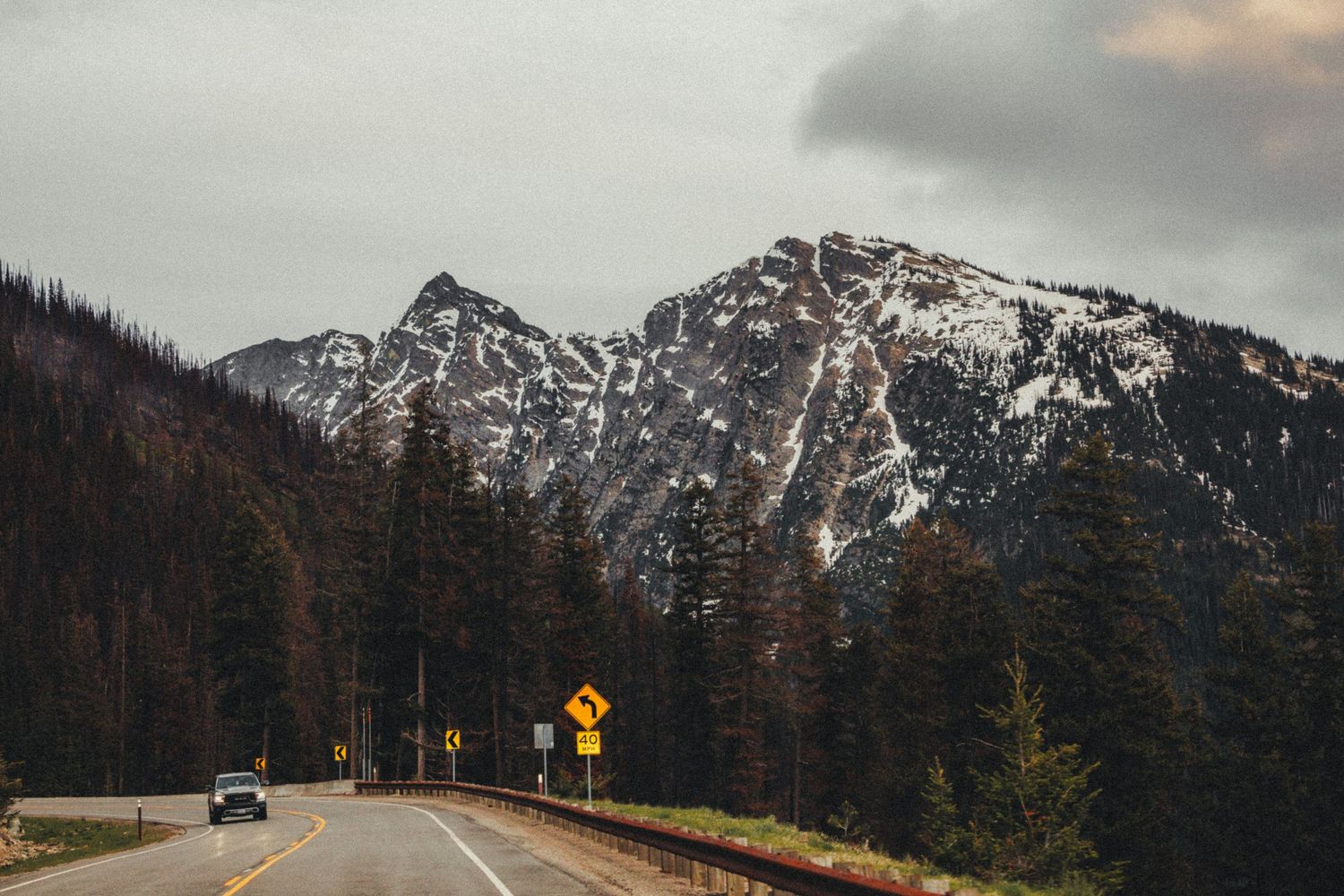Mountain highway road with snow-capped peaks, pine trees, and yellow warning signs under cloudy sky.