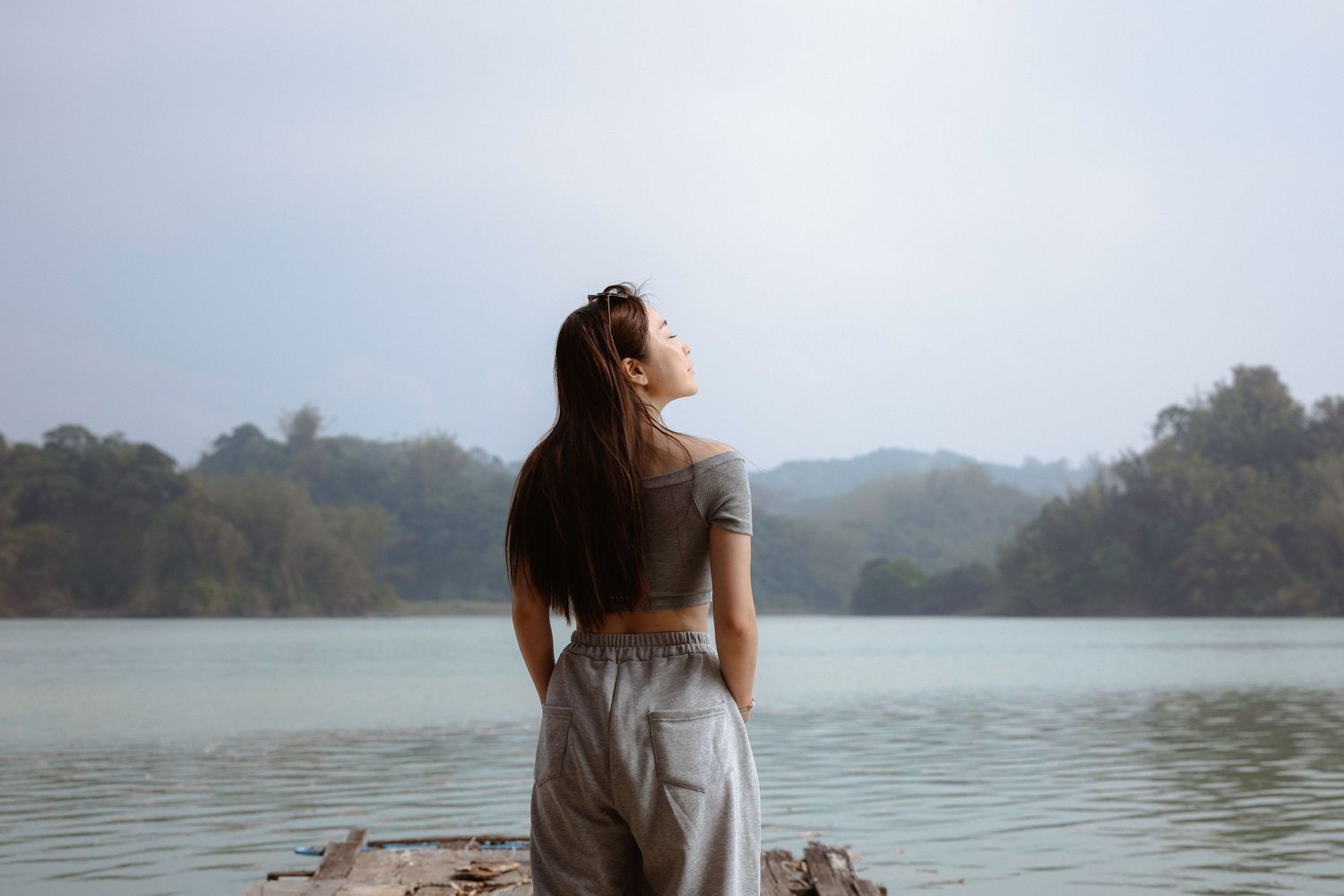 A young woman with a ponytail stands by a calm lake surrounded by misty green mountains.