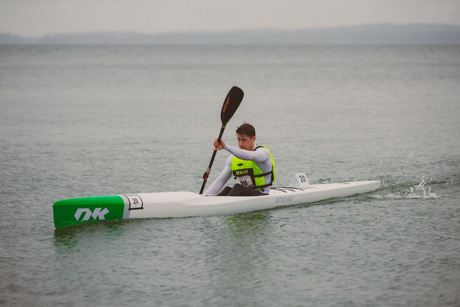 Young athlete paddles a white OK kayak on calm grey water during a kayaking race event.