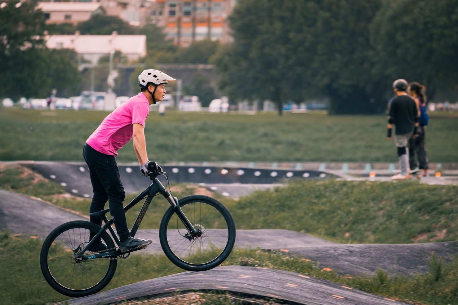 Female cyclist in pink shirt and white helmet riding a black bike on an outdoor pump track.