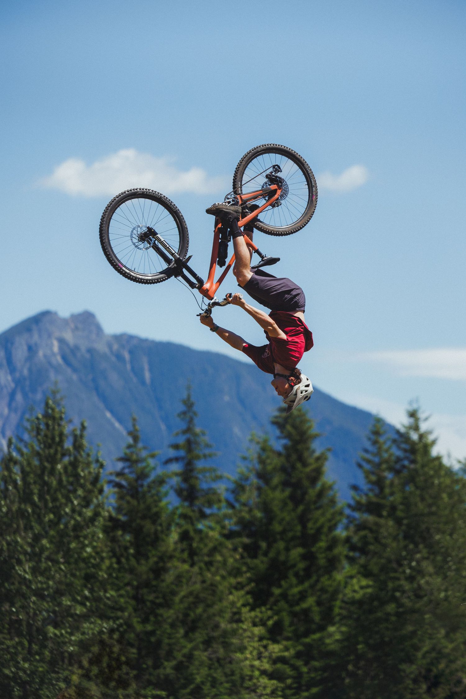 Extreme mountain biker performs an inverted backflip trick high above pine trees with a mountain backdrop.