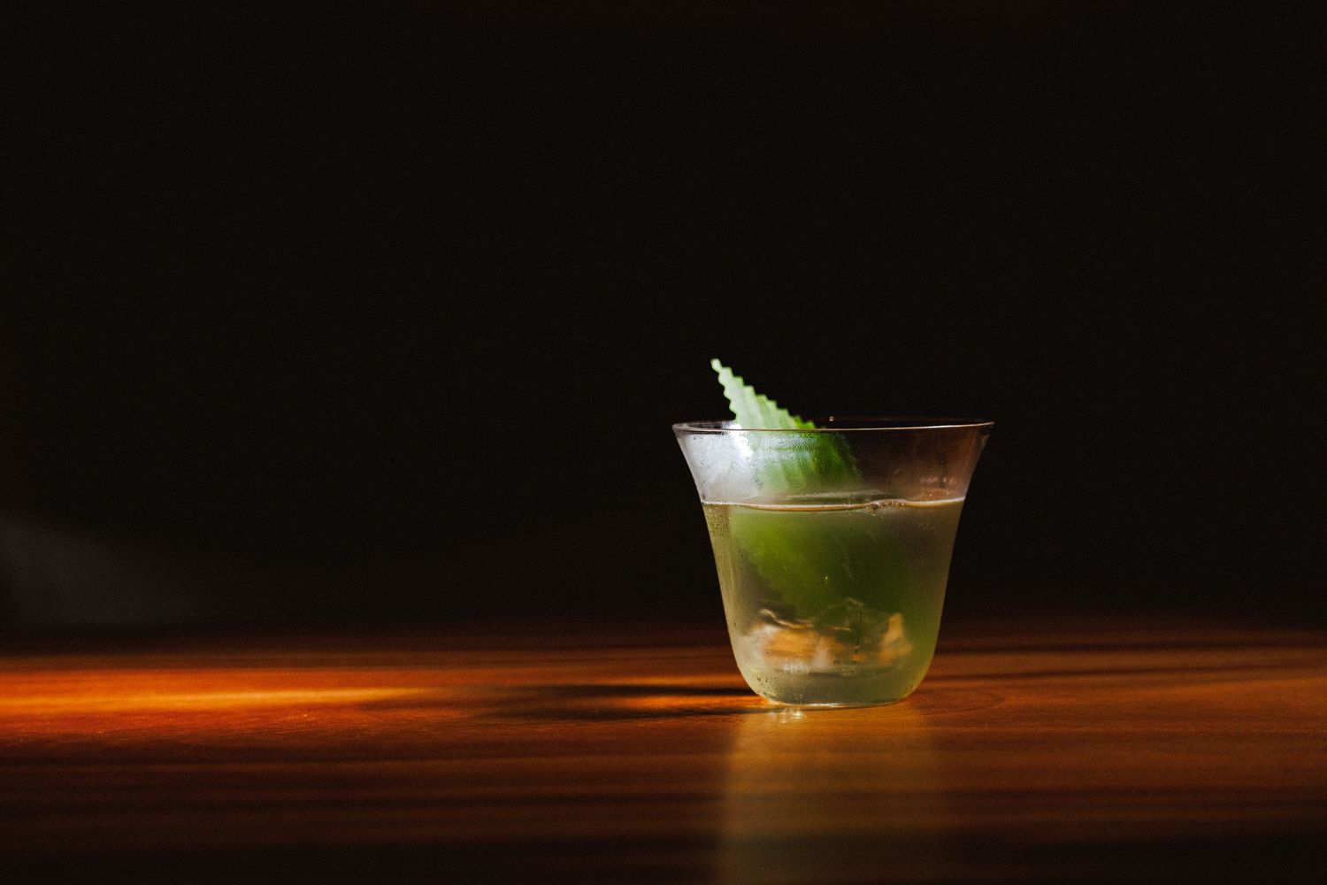 Green herb cocktail in crystal glass with fresh mint garnish on dark wooden bar surface.