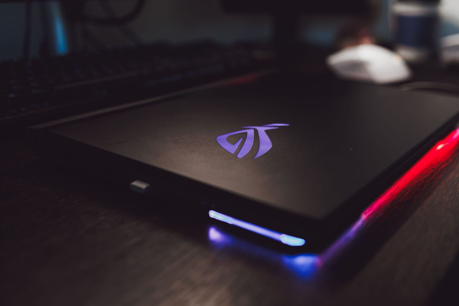ASUS ROG gaming laptop with glowing RGB lights and purple logo on dark desk setup.