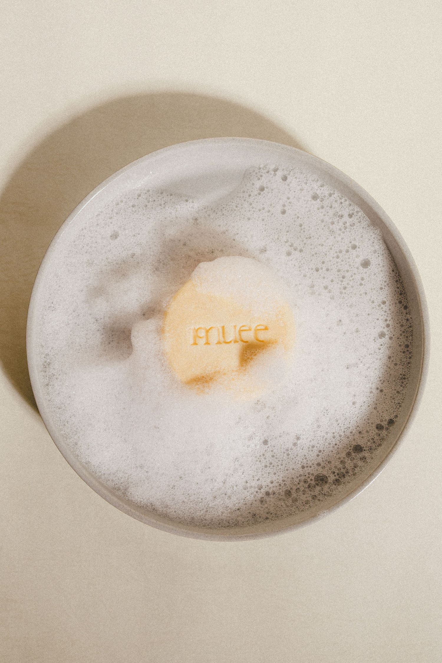 Muee branded bar soap floating in a bowl of white foam bubbles on a beige surface.