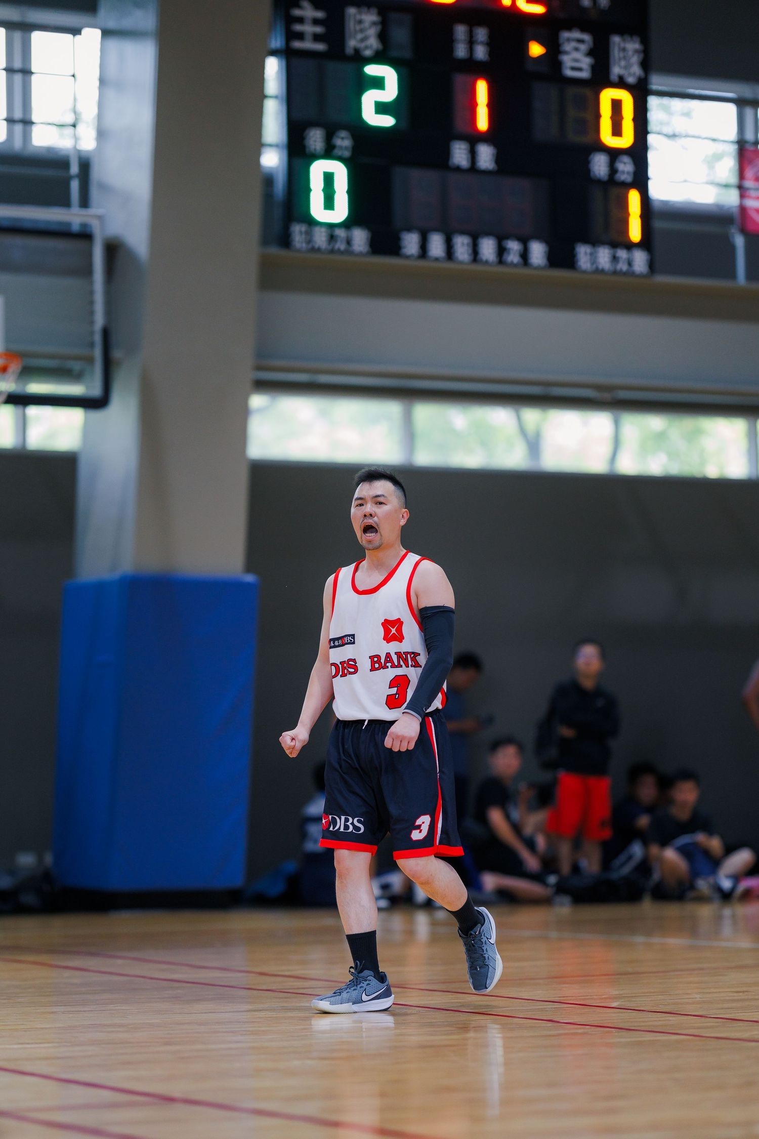 Basketball player wearing DBS jersey number 3 celebrating with fist pump in indoor court with scoreboard.