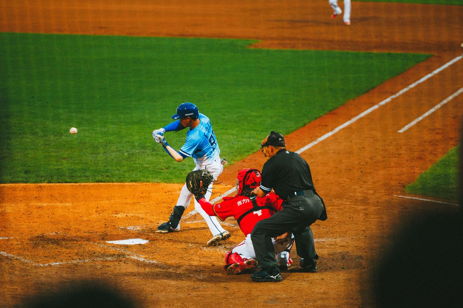 Baseball batter swings at a pitch while the catcher and umpire crouch behind home plate under stadium lights.