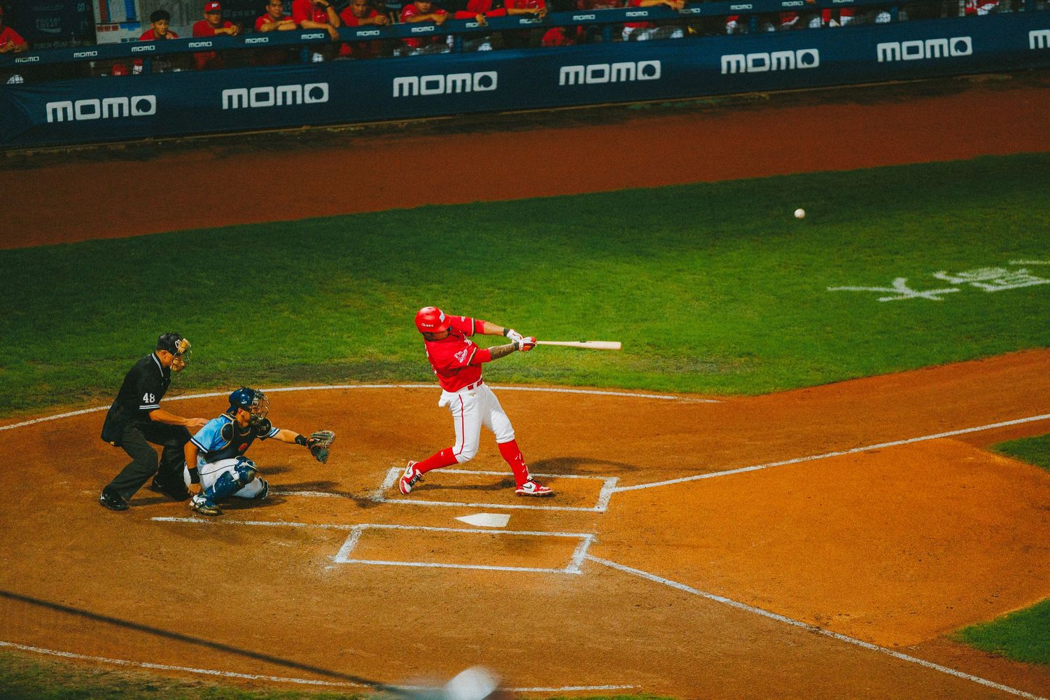 Baseball batter in red uniform swinging at a pitch during a night game with momo advertising boards behind.
