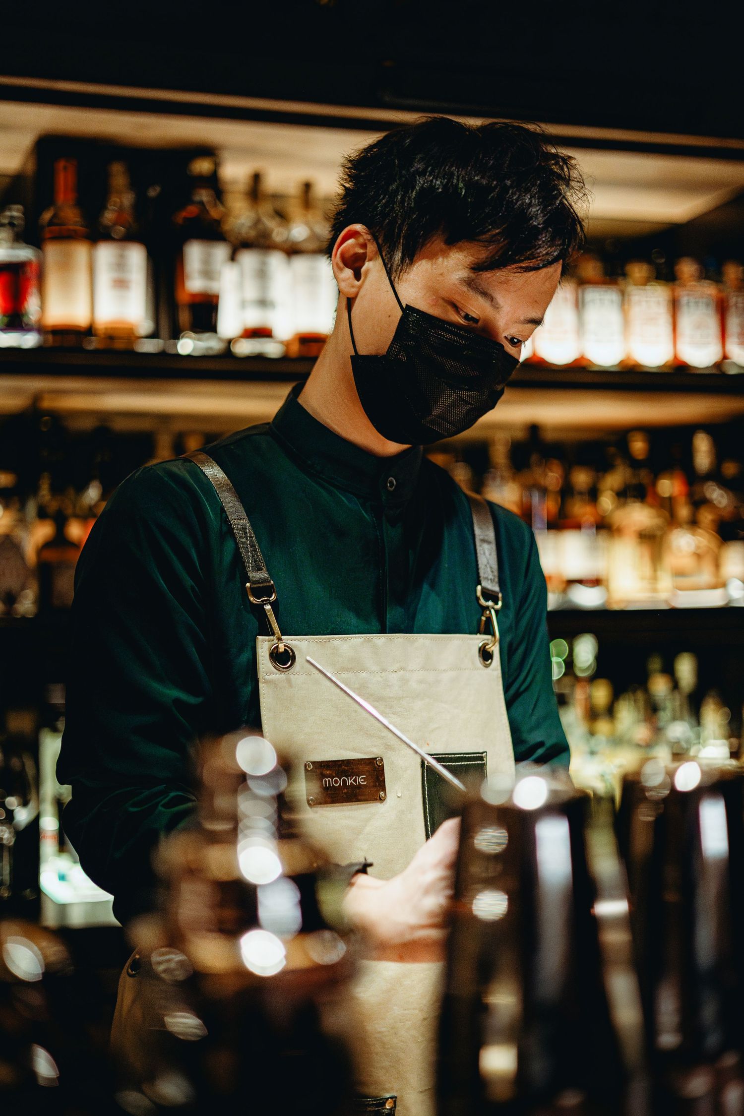Asian male bartender wearing black mask and apron preparing drinks in dimly lit cocktail bar.