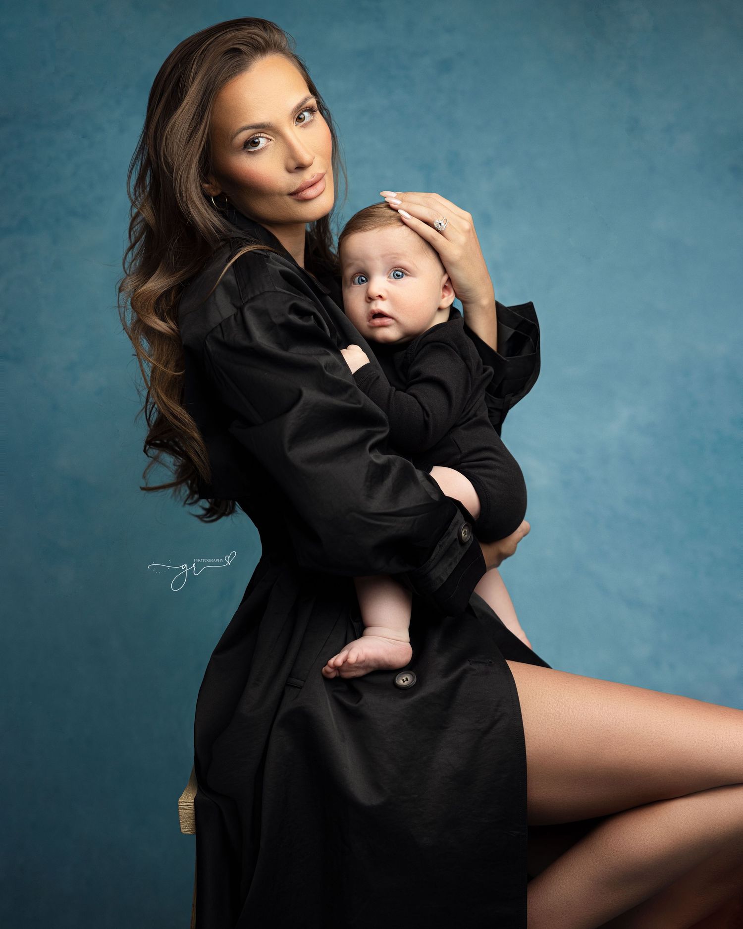 A woman in a black robe holds a blue-eyed baby in a studio portrait against a teal background.