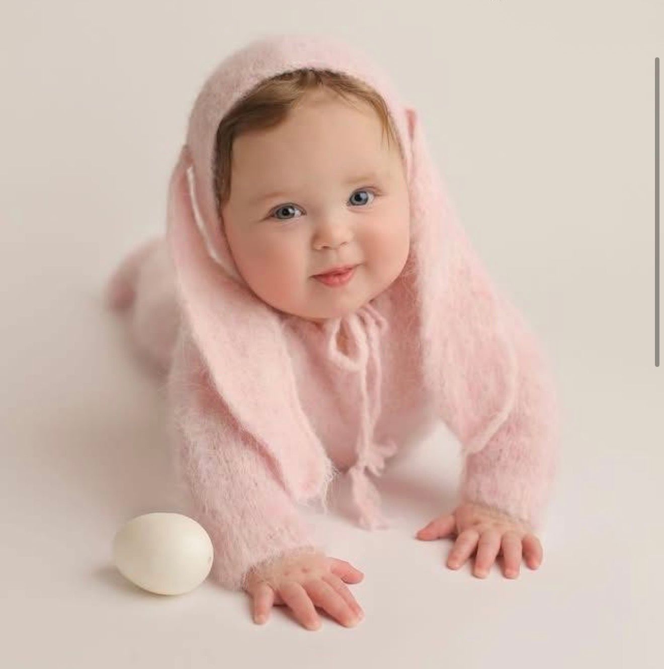 A baby in pink clothing crawls and reaches for white eggs on a light background.