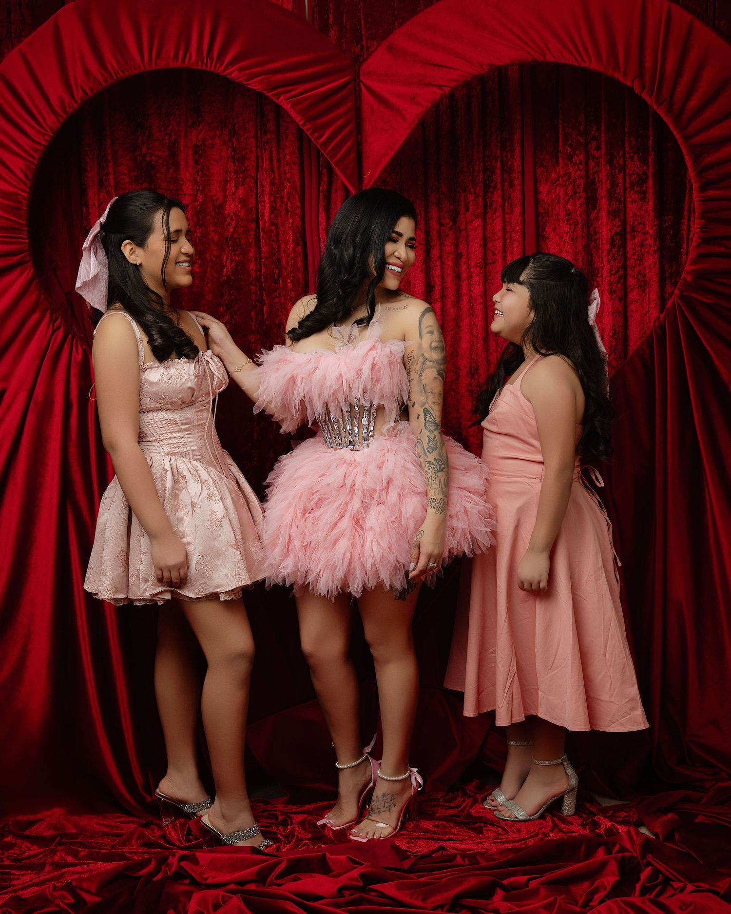 Three friends in pink dresses pose playfully against a dramatic red velvet curtain backdrop during a photo session.
