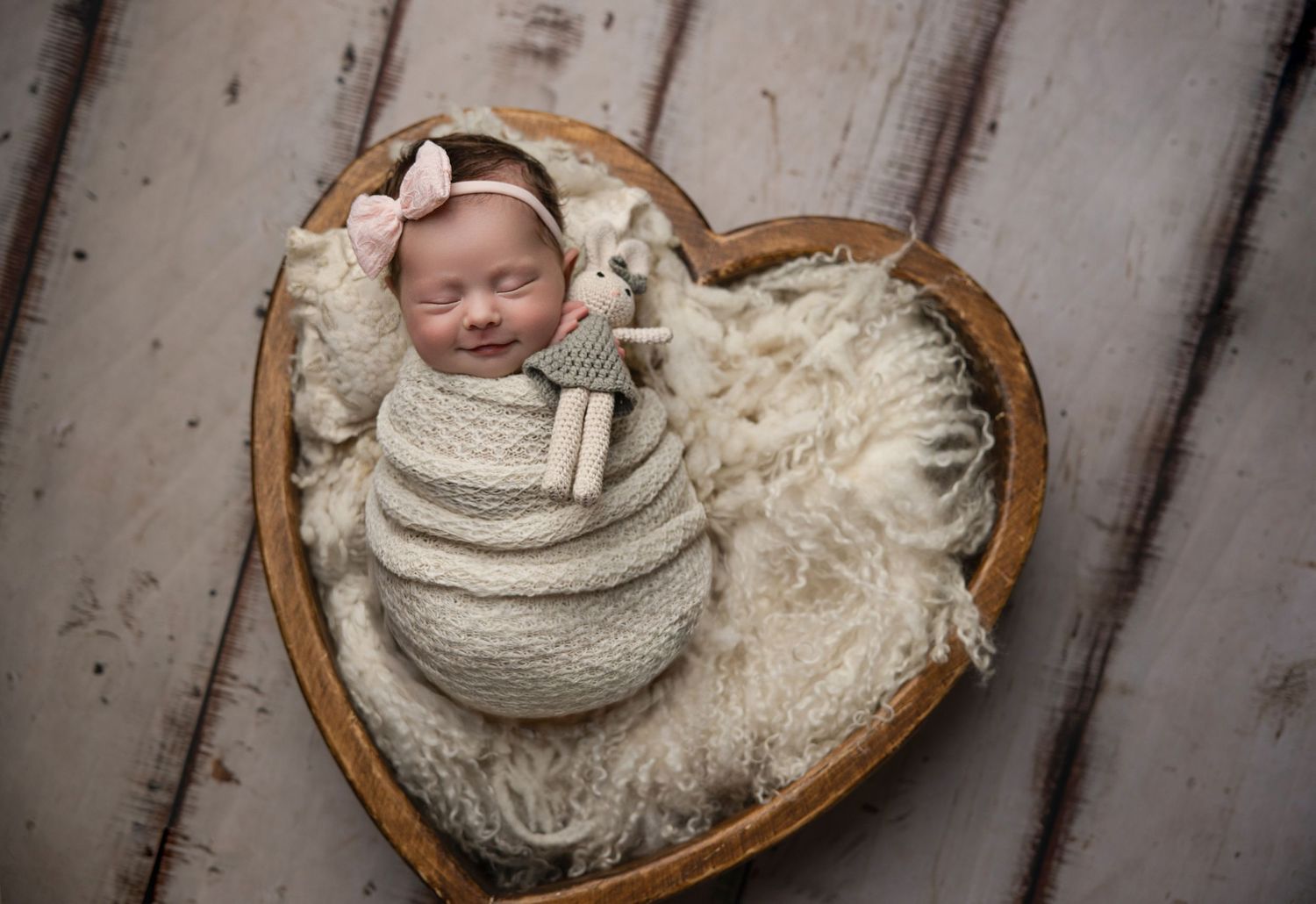 Smiling newborn baby swaddled in cream wrap, nestled in heart-shaped wooden bowl with fluffy blanket and bunny toy.