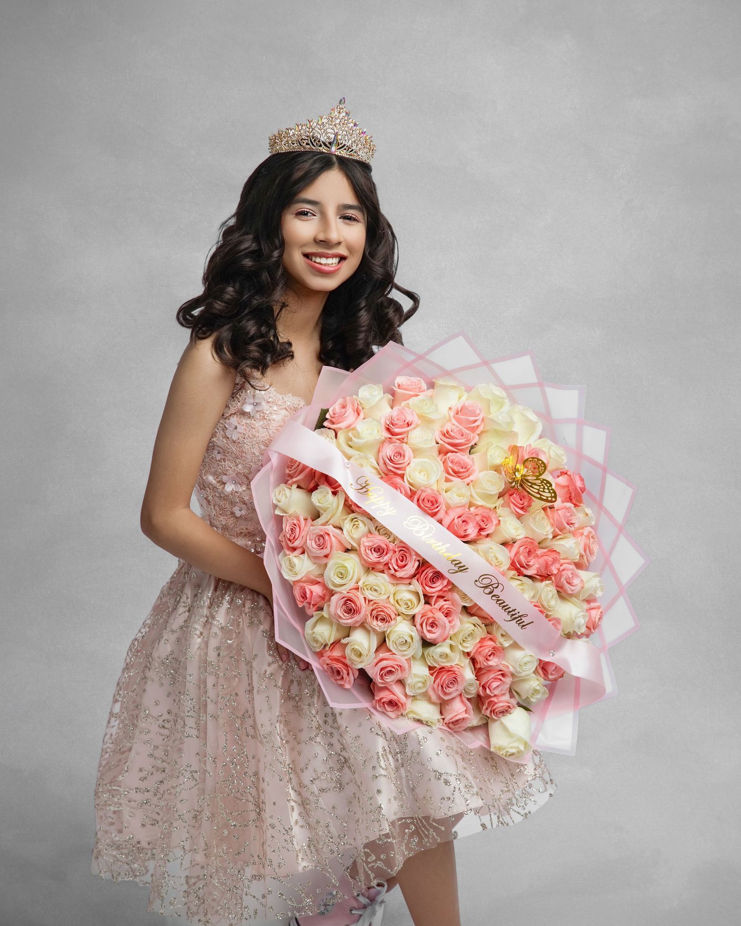 A person in a pink lace dress and tiara holds a large bouquet of pink and cream roses wrapped in delicate paper.